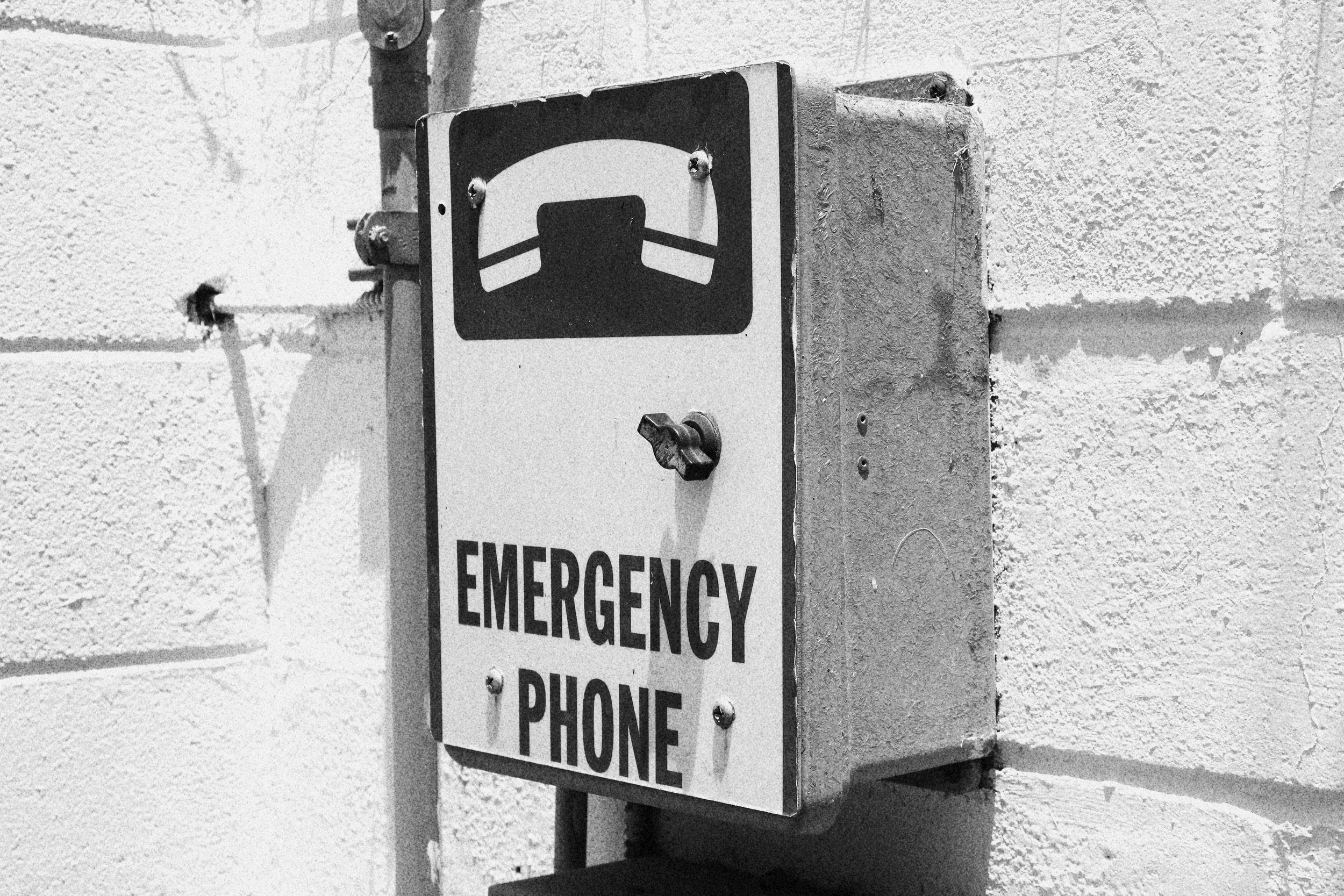 Black and white photo of an outdoor emergency phone box mounted on a brick wall, with a pictogram of a telephone receiver and the words "EMERGENCY PHONE" beneath.
