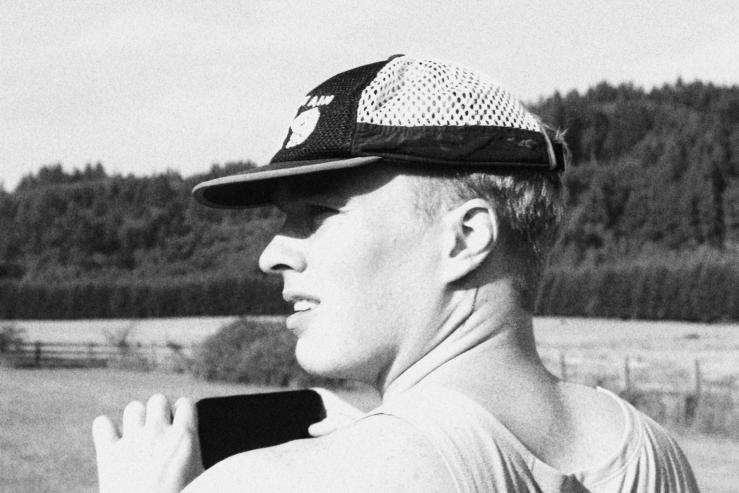 A black and white photo of a person wearing a cap, looking to the side, outdoors with trees and fencing in the background.