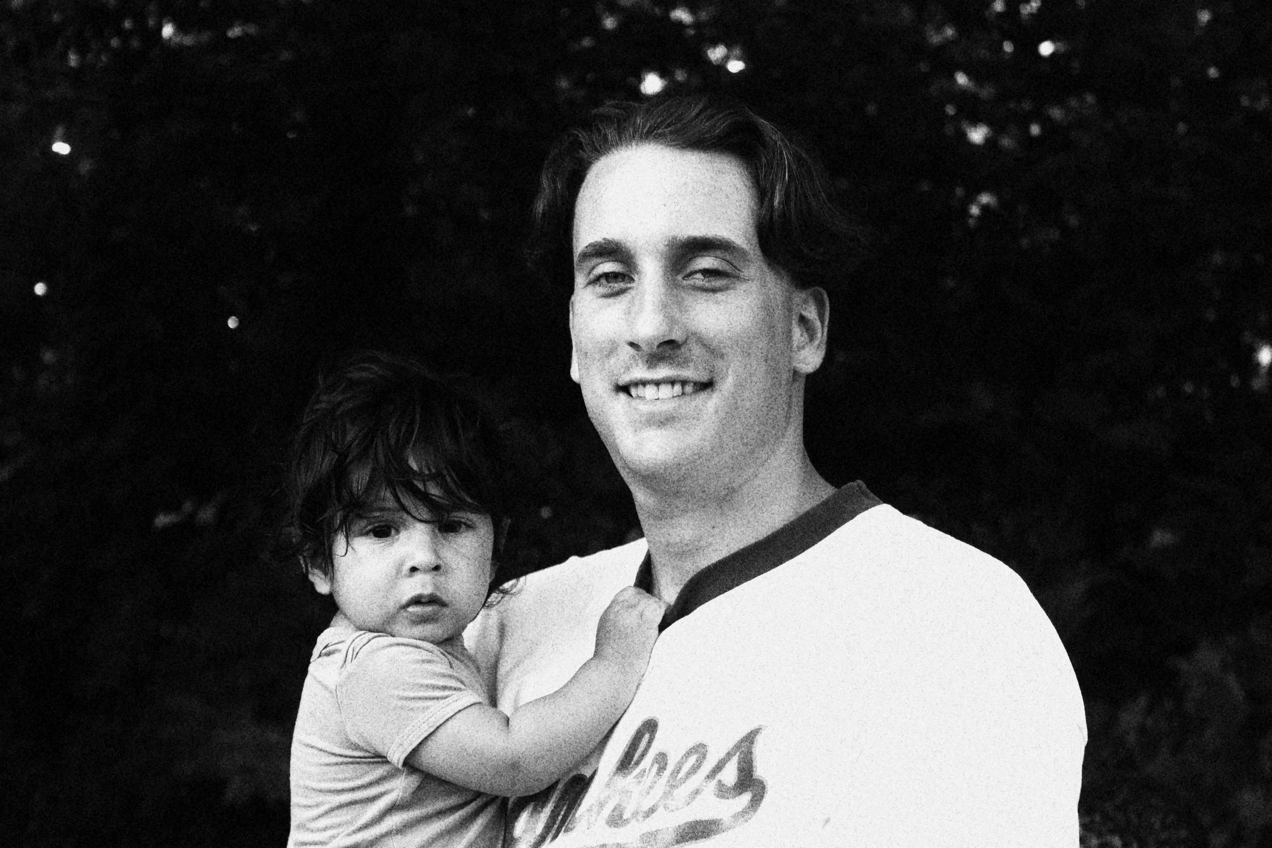 A man holding a young child in his arms outdoors at dusk, both facing the camera with a dark, blurred natural background.
