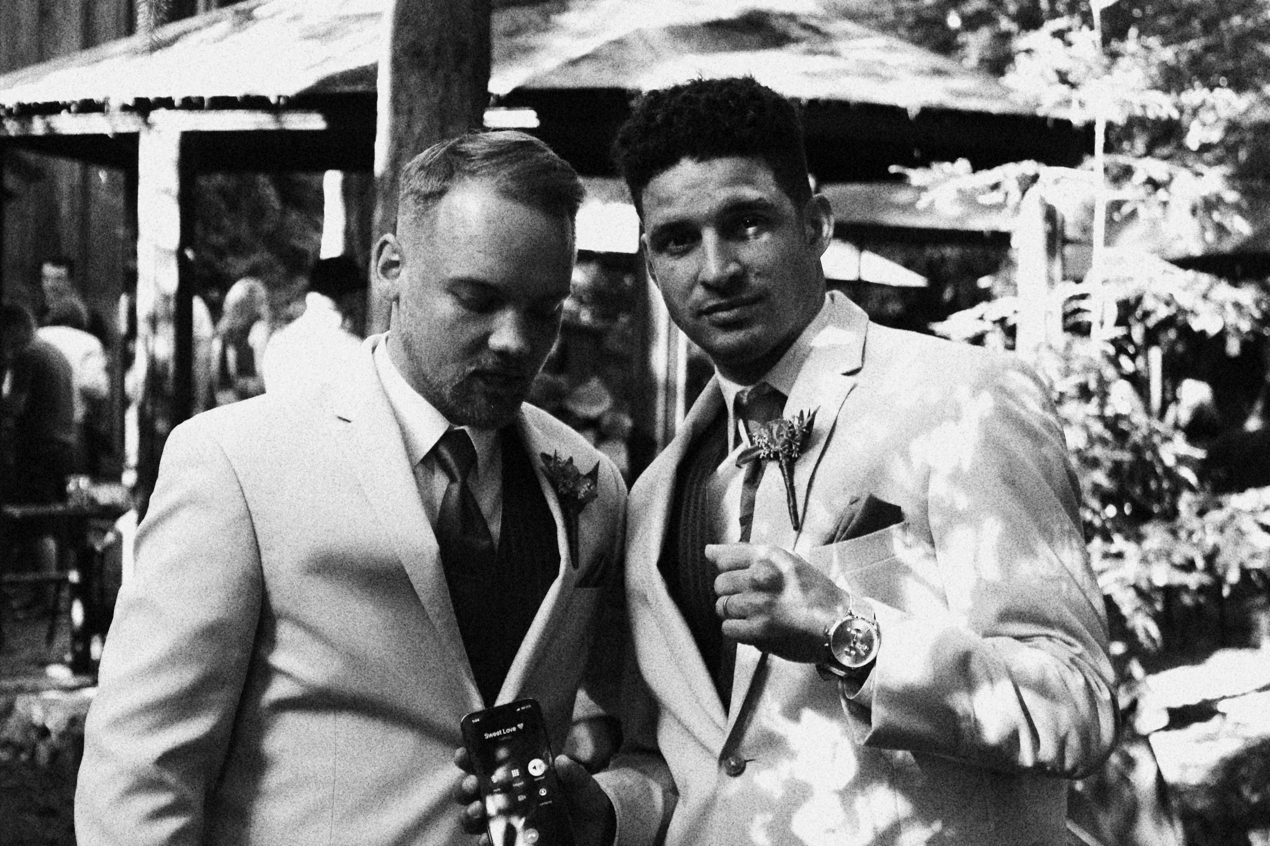 Two men dressed in tuxedos at an outdoor event, one looking at his phone and the other pointing at the camera, with trees and people in the background.