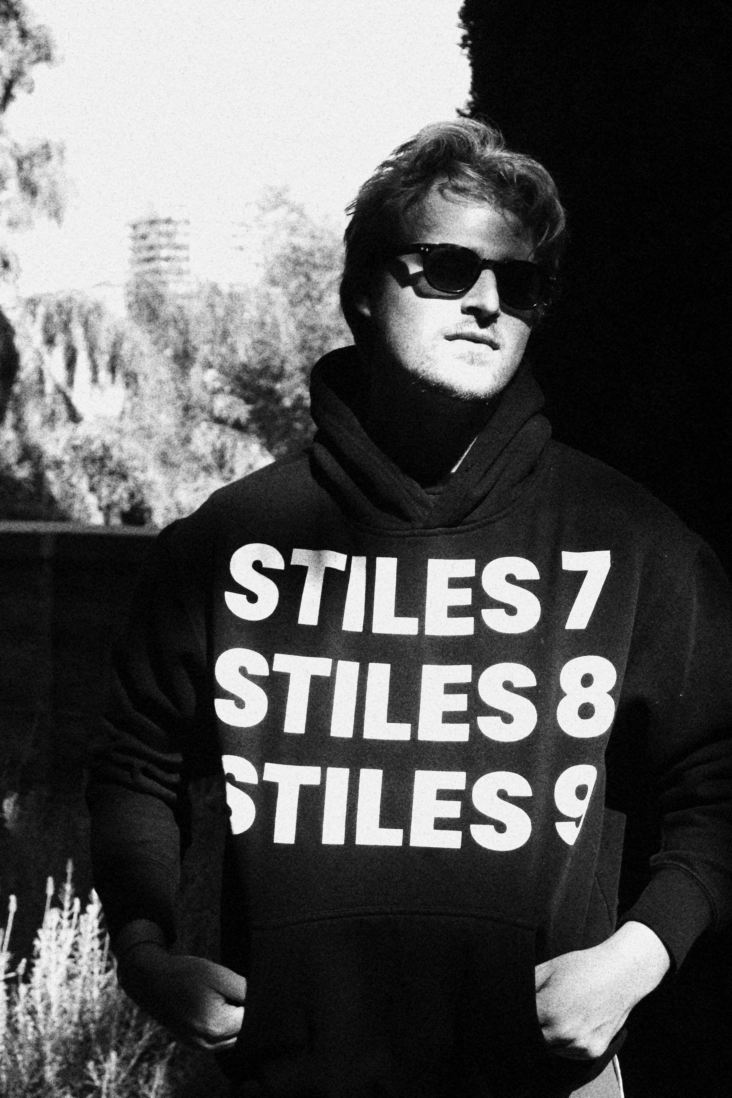Young man wearing sunglasses and a black hoodie with large text reading 'STILES 7, STILES 8, STILES 9' standing outdoors in sunlight.