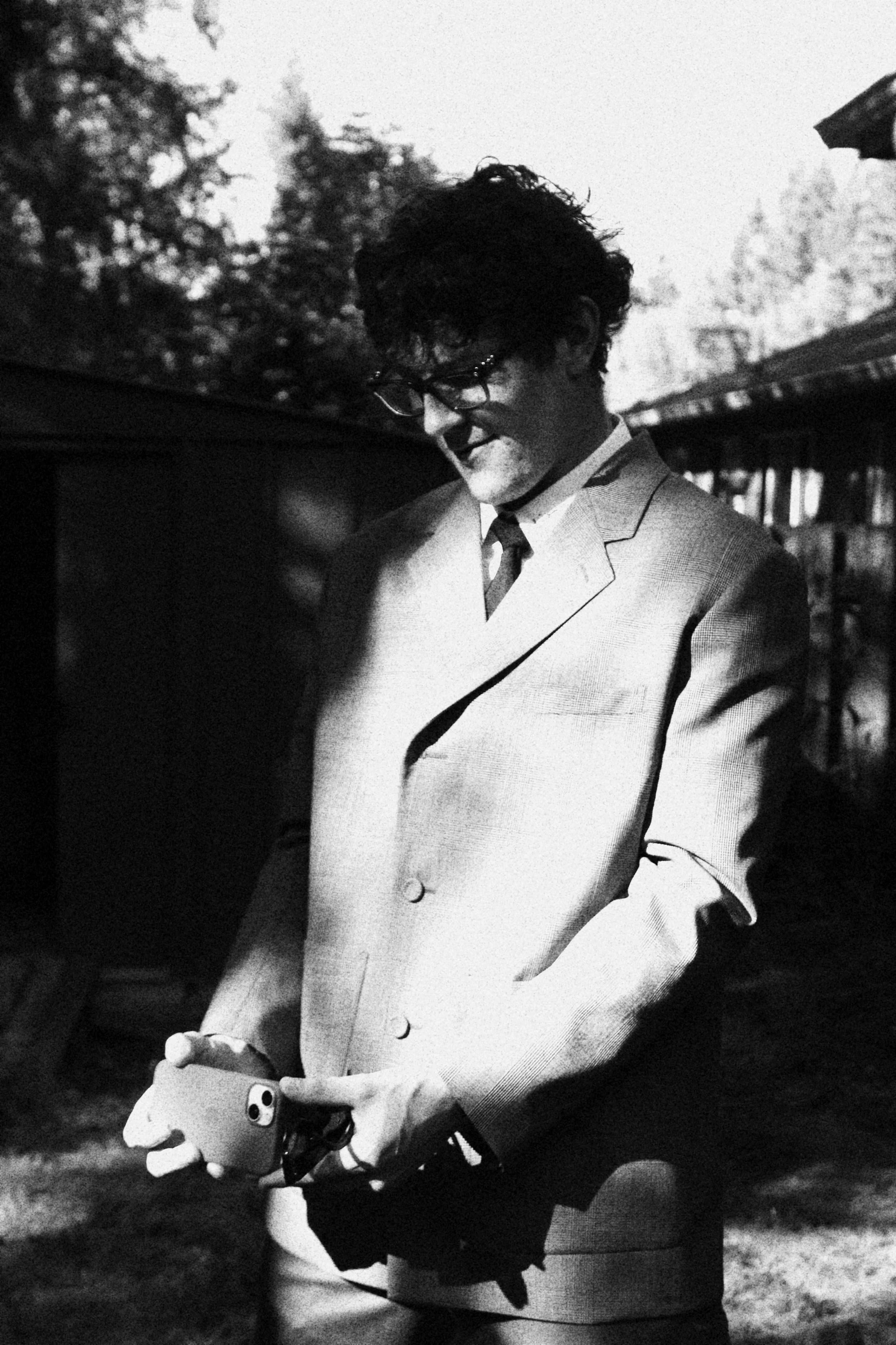 A man in a suit and glasses looking at his phone outdoors, with trees and a building in the background in black and white.