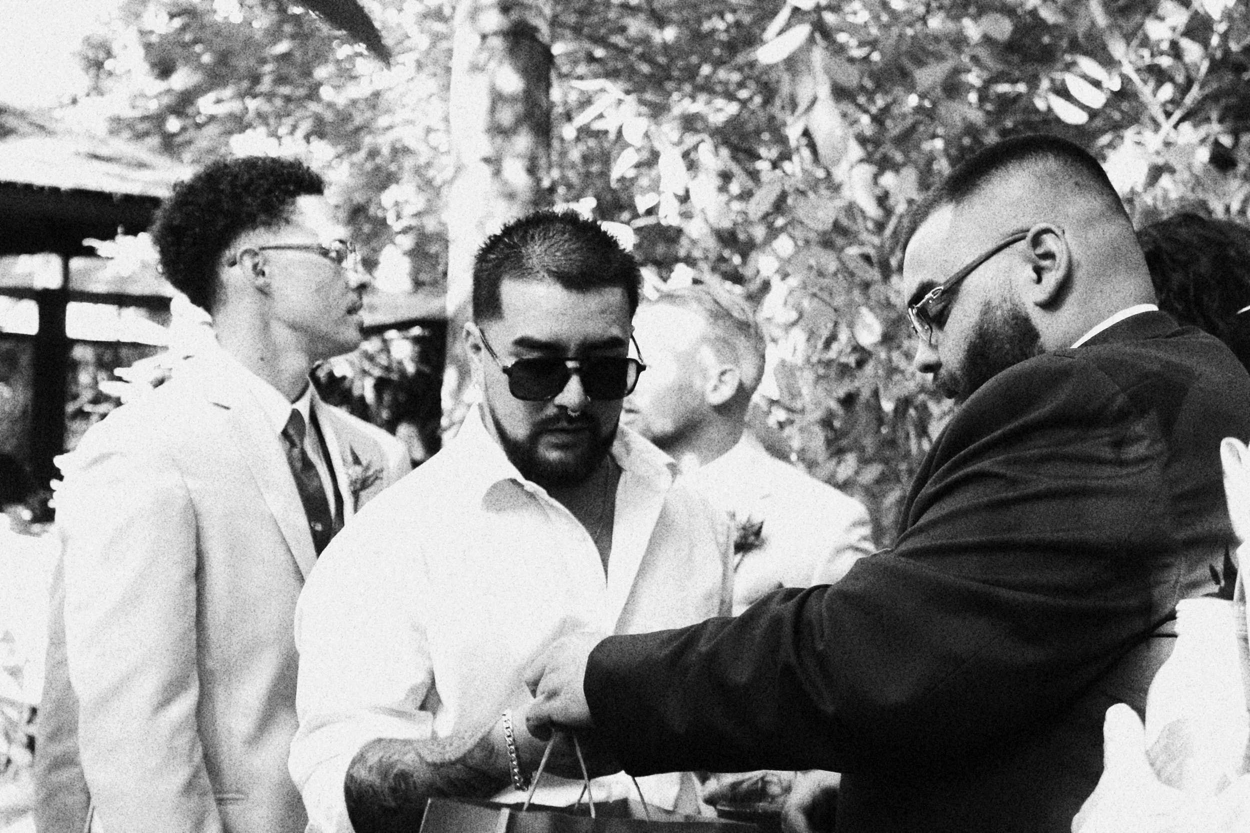 Group of four men in formal attire, one in a black suit, appearing to exchange an item or gesture, outdoors with trees in the background.