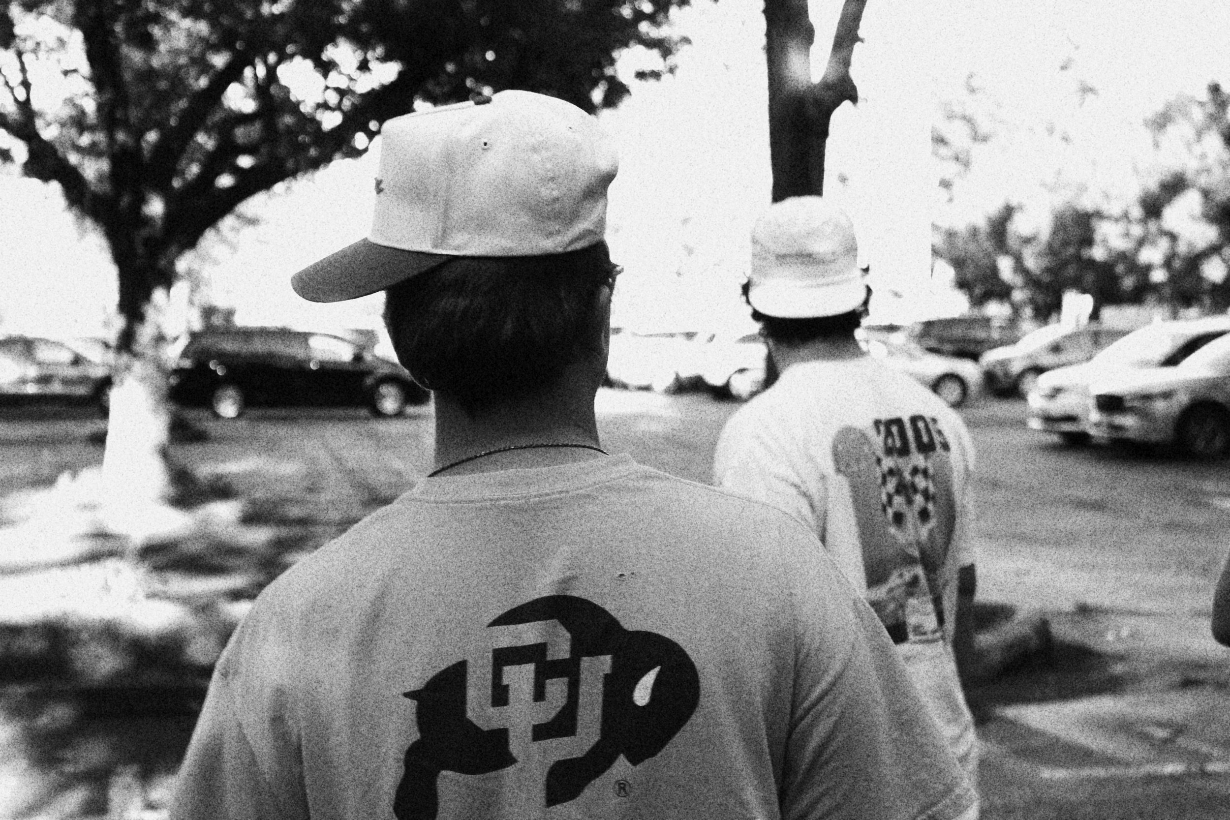 Two young men are walking outdoors in a parking lot with trees, wearing t-shirts and caps. The photo is in black and white.