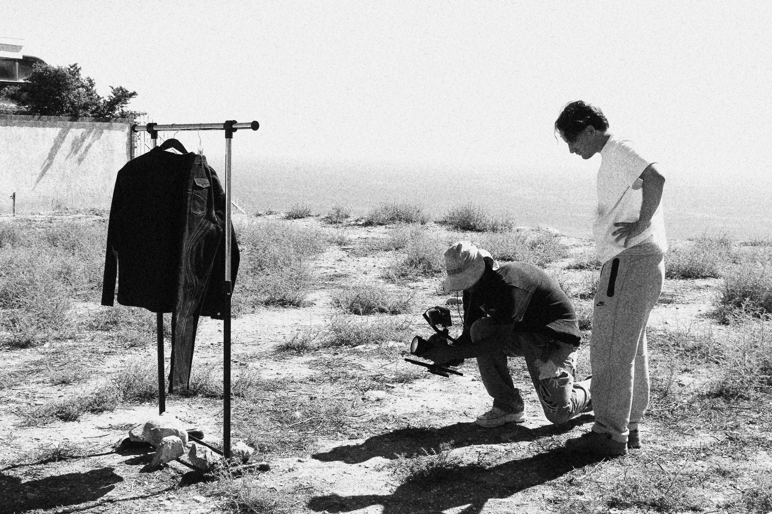 Two men filming outdoors with a coat hanging on a stand in a desert-like area with sparse bushes, under a bright sun.