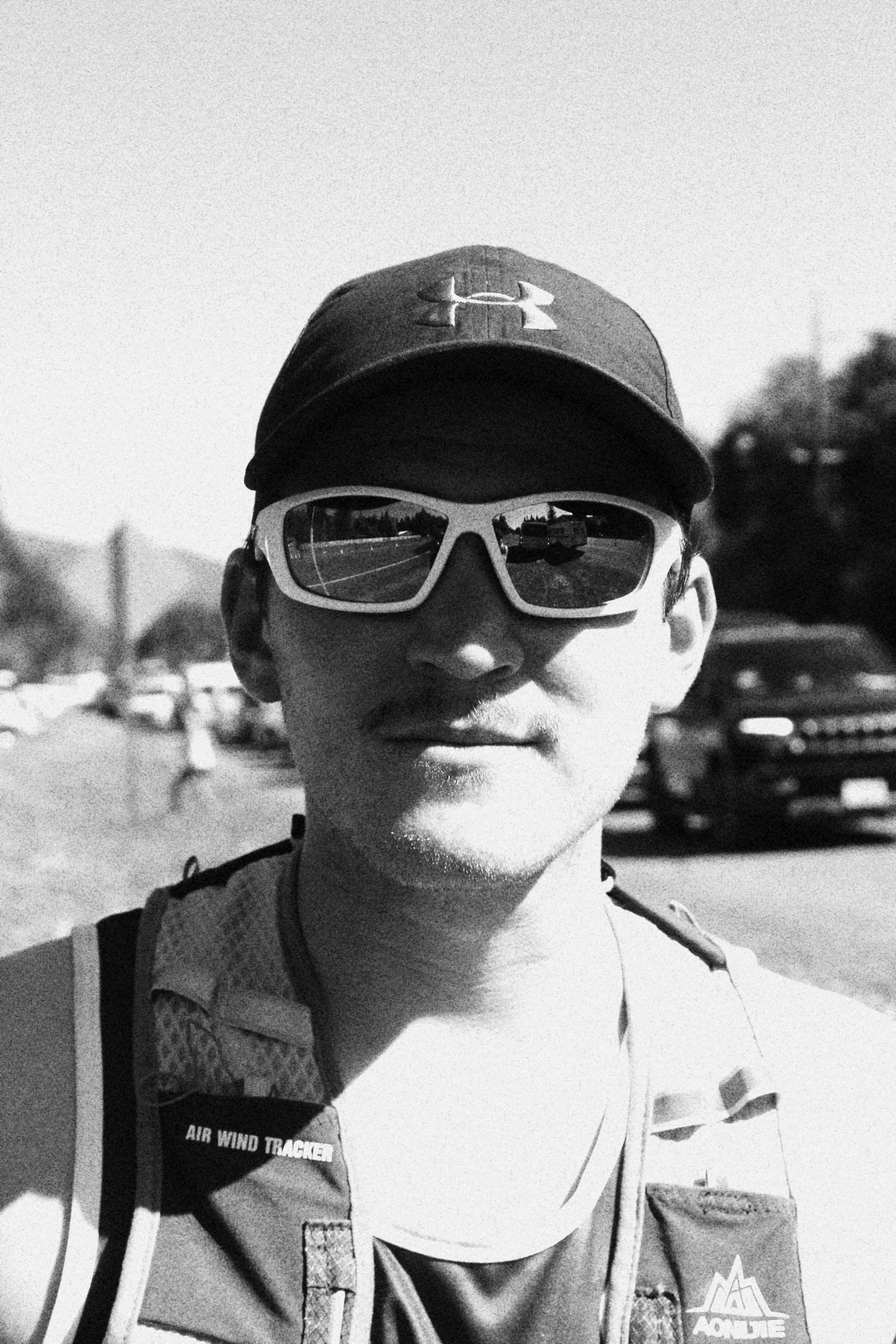 A person wearing a baseball cap, sports sunglasses, and athletic gear outdoors on a sunny day, with cars and trees in the background.