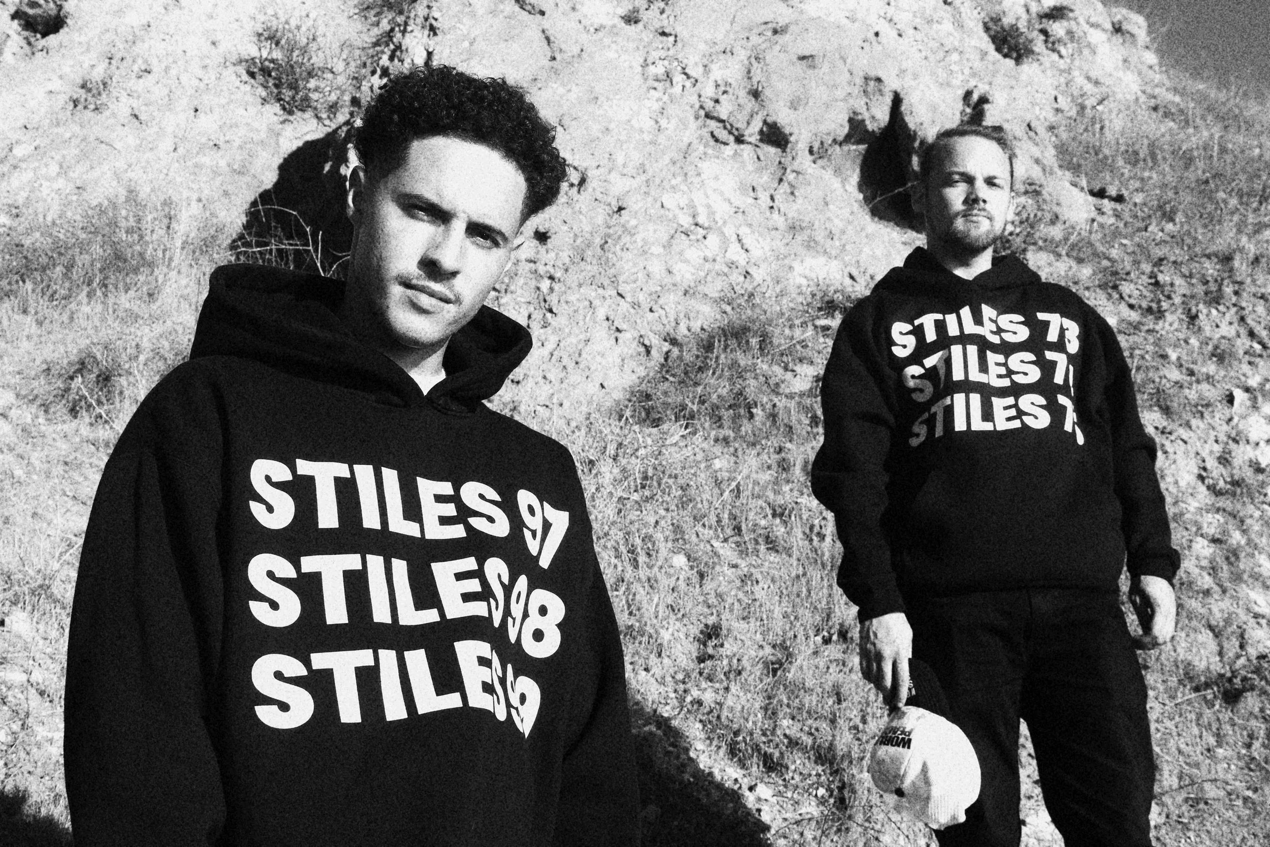 Two men standing outdoors in front of a rocky hillside, both wearing black hoodies with the word 'STILES' and a number printed multiple times on the front. One is looking slightly left, the other slightly right, with serious expressions.