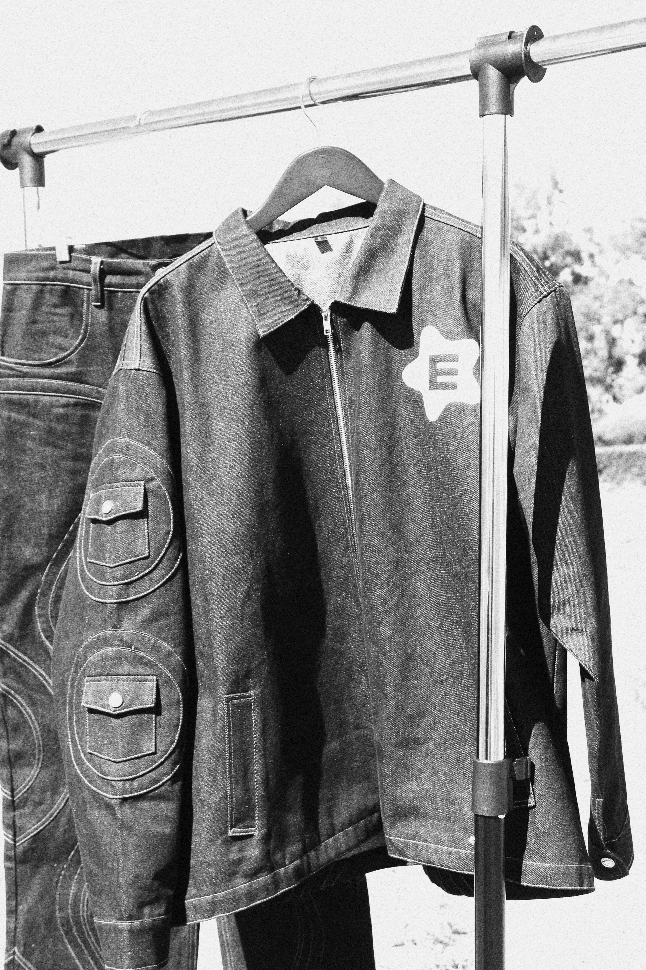 Black and white photo of clothing hanging on a rack, including a jacket with a star-shaped patch featuring the letter 'E' and a pair of jeans.