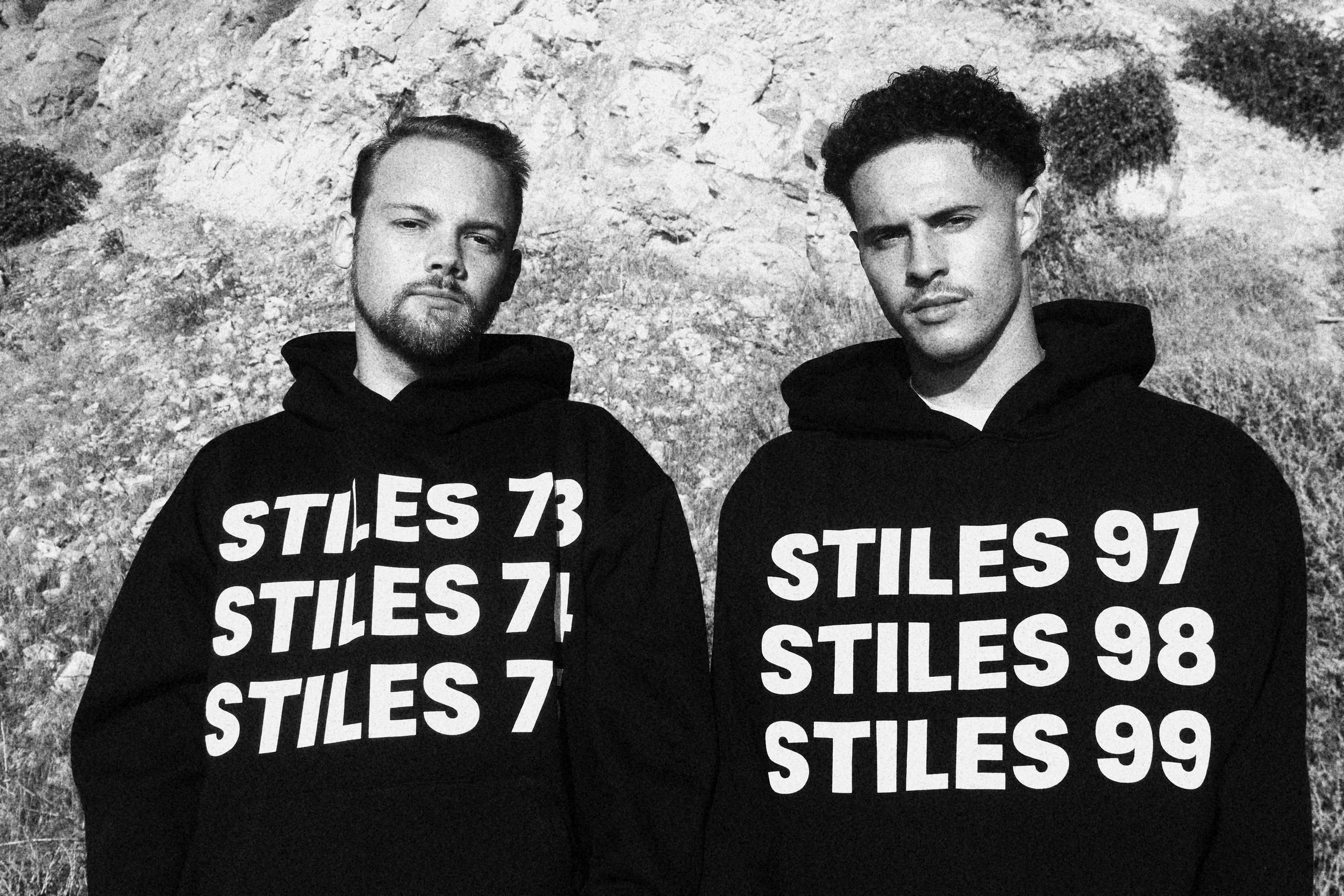 Black and white photograph of two young men standing outdoors against a rocky background, both wearing black hoodies with bold white text listing numbers and the word playing 'STILES'.