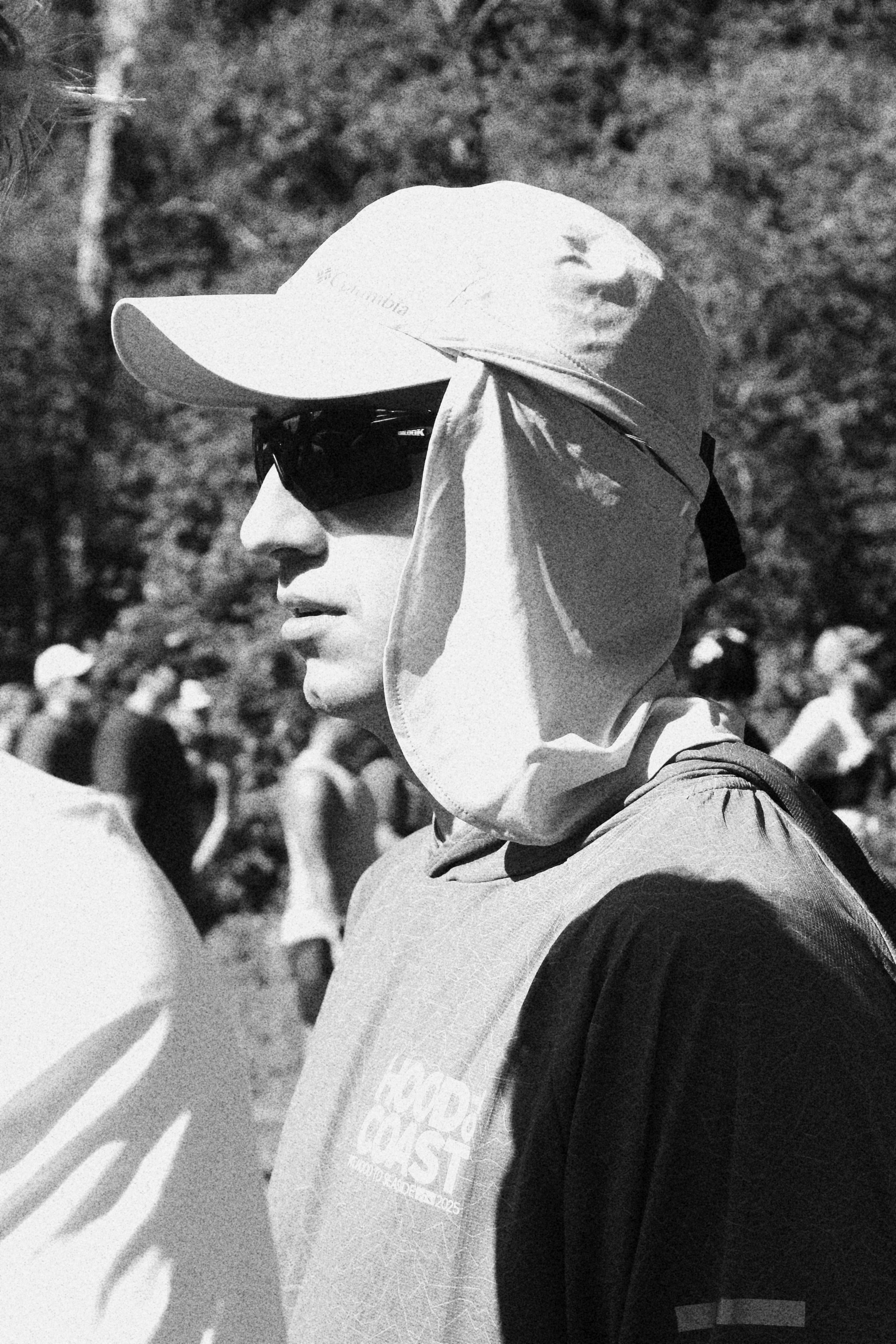 A person wearing a baseball cap, sunglasses, and a hoodie, participating in an outdoor event with other people in the background.