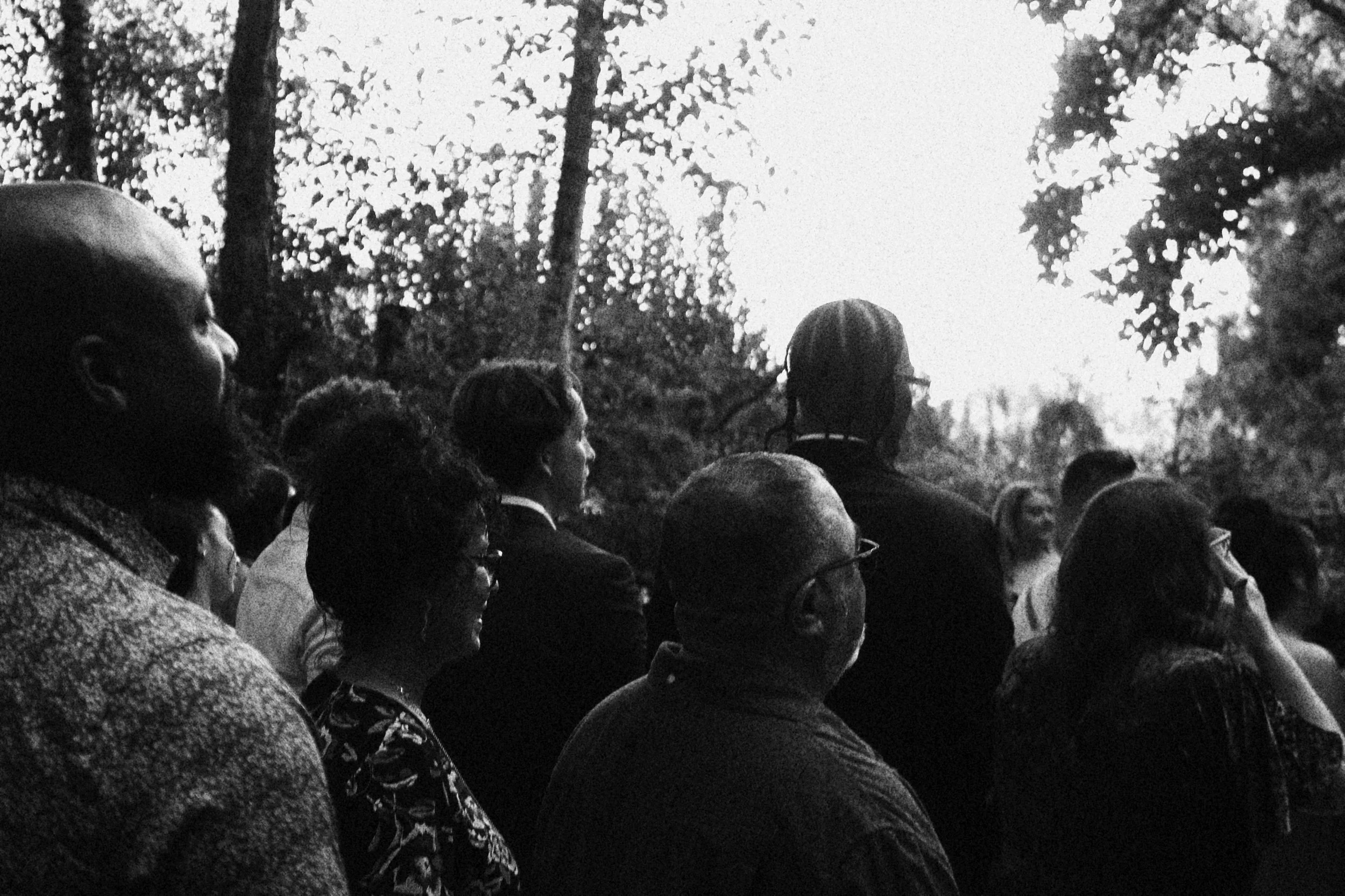 A group of diverse people gathered outdoors, facing a bright sky, in a wooded area, possibly attending an event or listening to a speaker.
