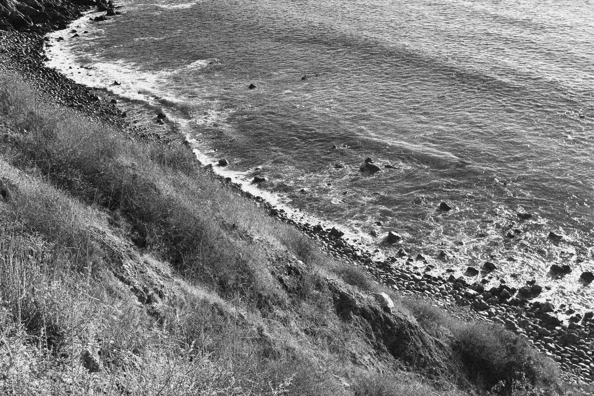 Black and white photograph of a rocky shoreline with waves crashing against the coast and sparse vegetation on the hillside.