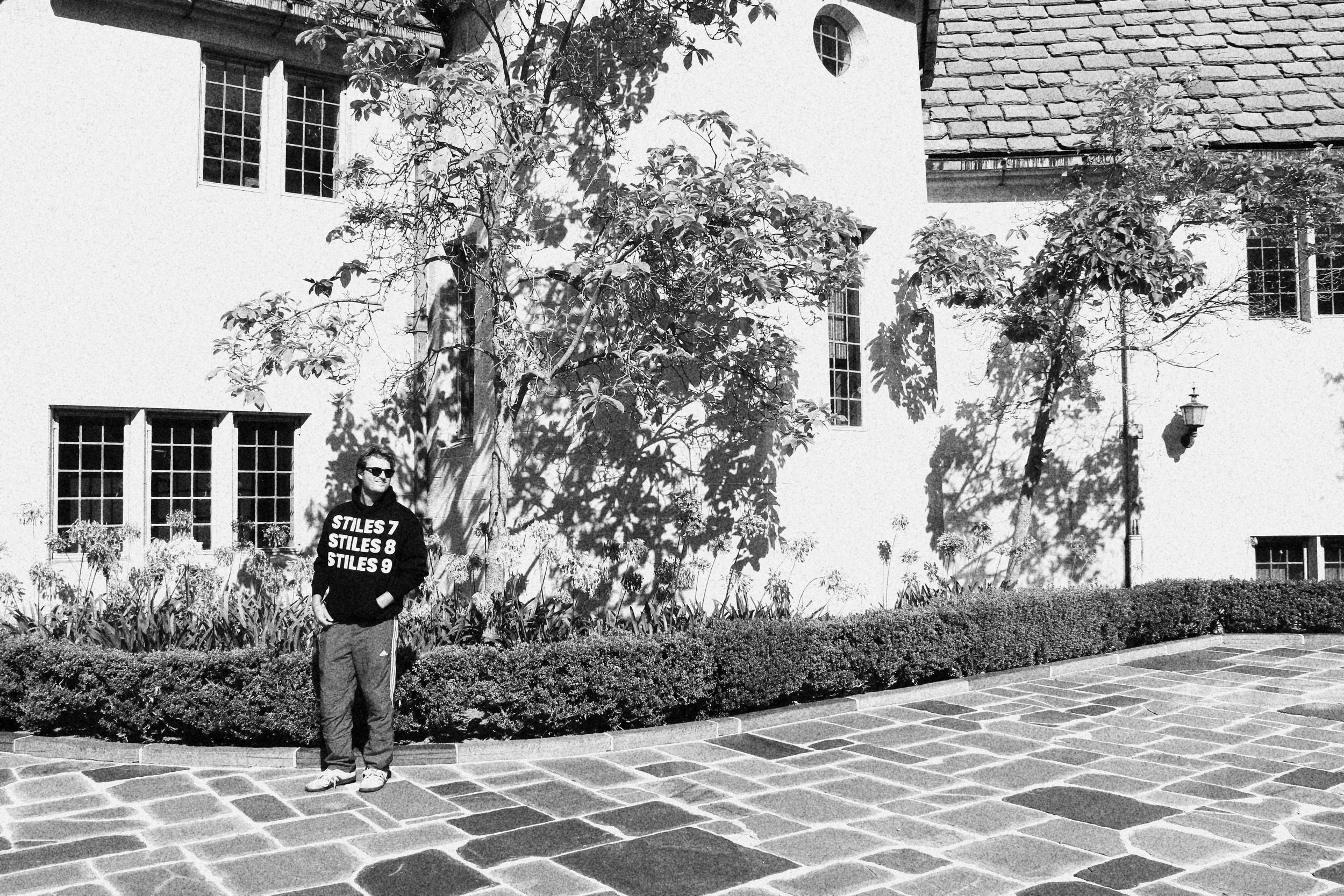 A person standing outdoors in front of a white building with multiple windows and a hedge, wearing sunglasses and a black hoodie with white text, in a black and white photo.