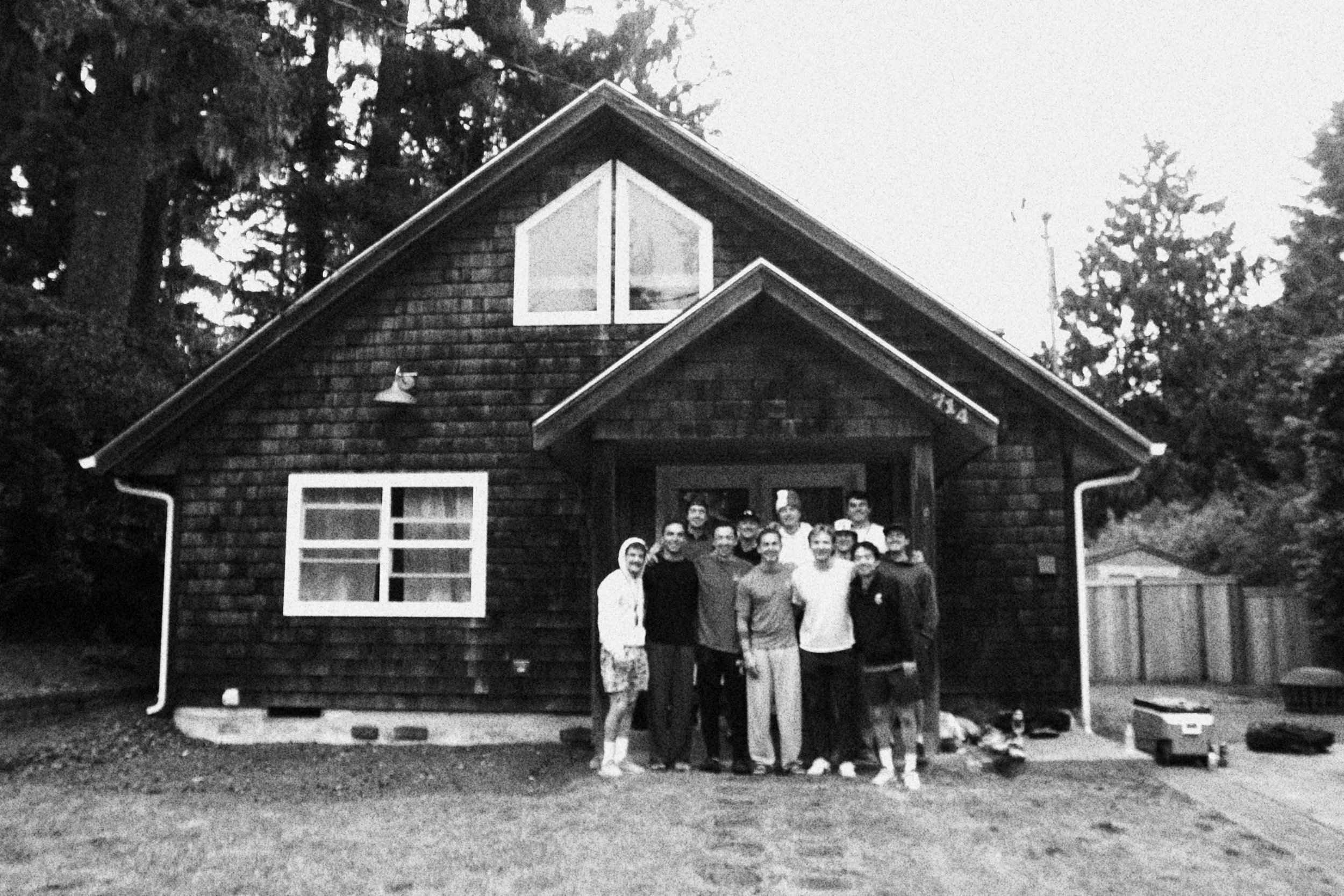 A group of people standing in front of a dark wooden house with a gabled roof and large windows, surrounded by trees in the background.