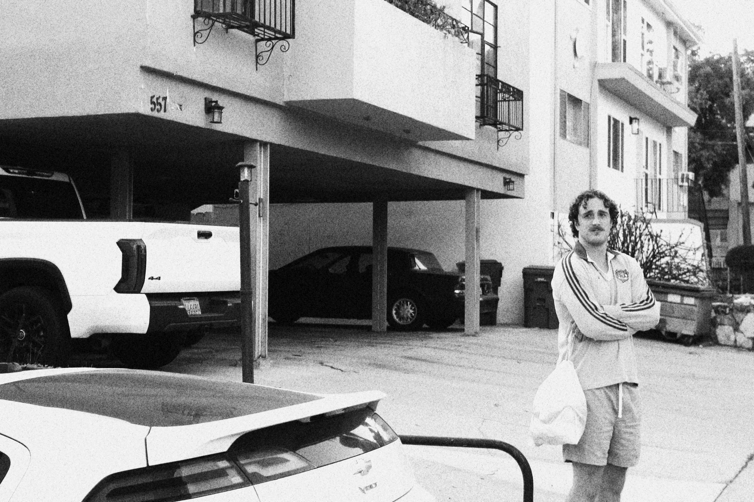 A man with curly hair and a mustache wearing a windbreaker jacket and shorts, standing on a sidewalk with arms crossed, in front of an apartment building with parked cars, trash bins, and balconies.
