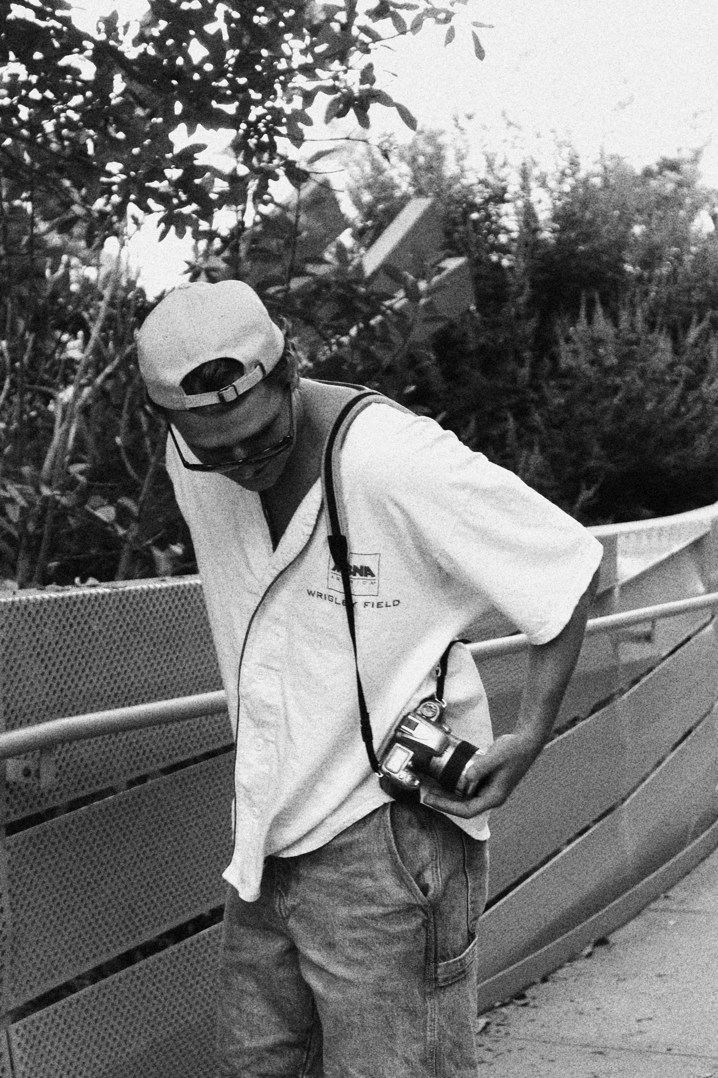 A young man wearing a cap backwards, sunglasses, a polo shirt with the logo 'WRIGLEY FIELD,' and jeans, holding a camera with a strap around his neck, standing on a bridge or walkway outdoors with trees and bushes in the background.