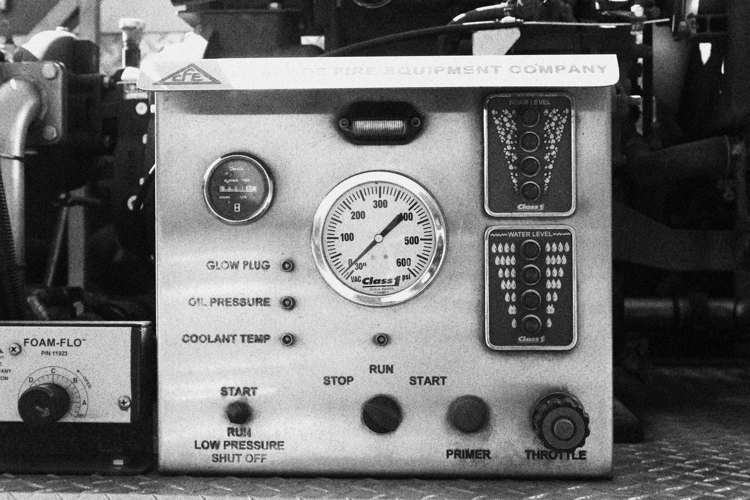 A black and white photograph of a mechanical control panel with gauges, dials, buttons, and indicators, labeled with various functions like oil pressure, coolant temperature, and water level.