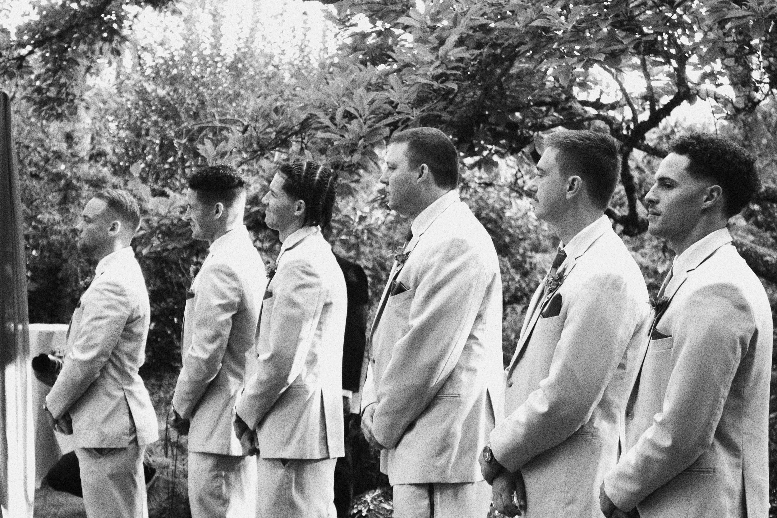 A black and white photo of six men in white suits standing in line outdoors, likely during a formal event or ceremony, with trees and foliage in the background.