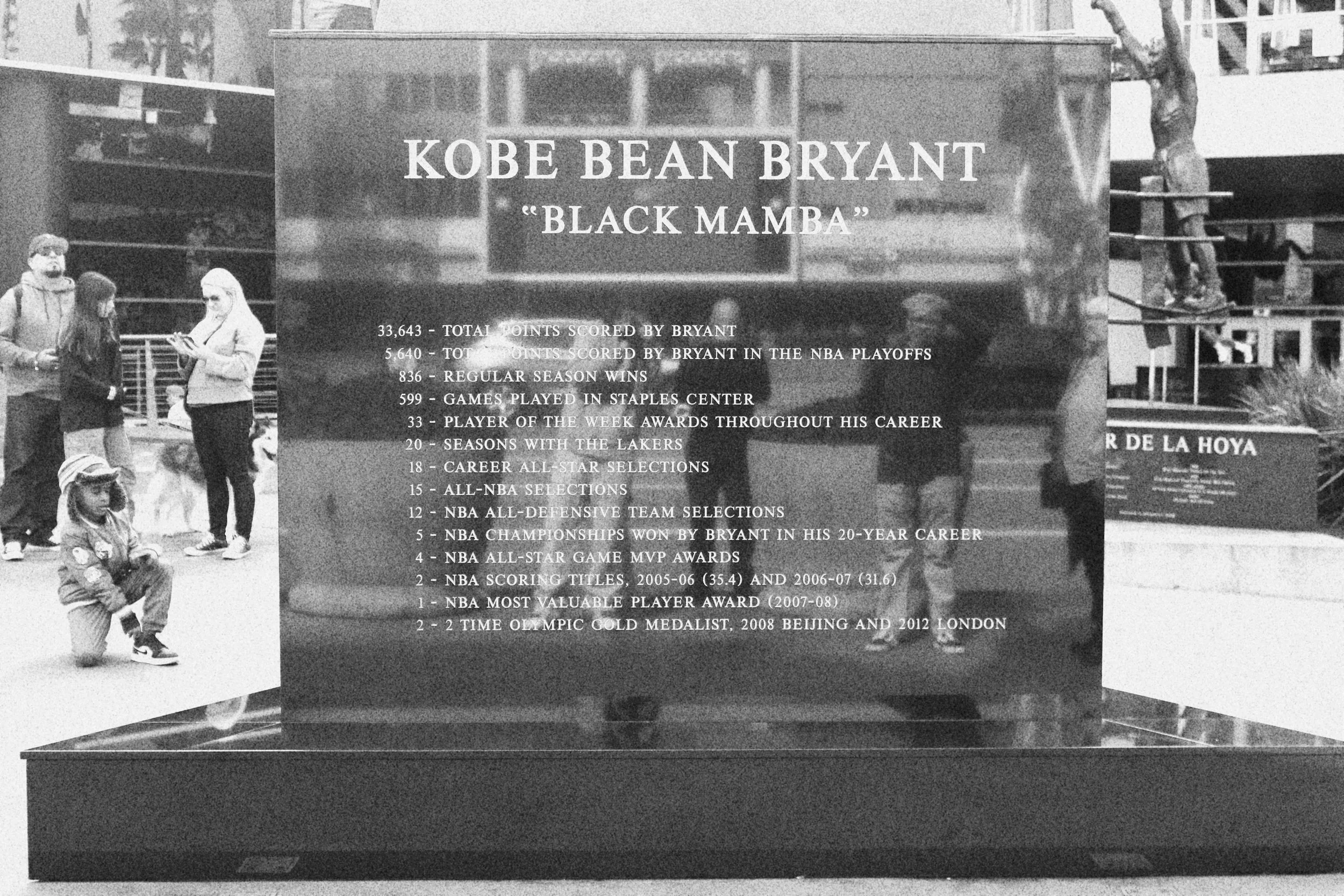 Plaque displaying Kobe Bryant's achievements and statistics, including points scored, MVP awards, and championships, with a background of people outside in a park or plaza.
