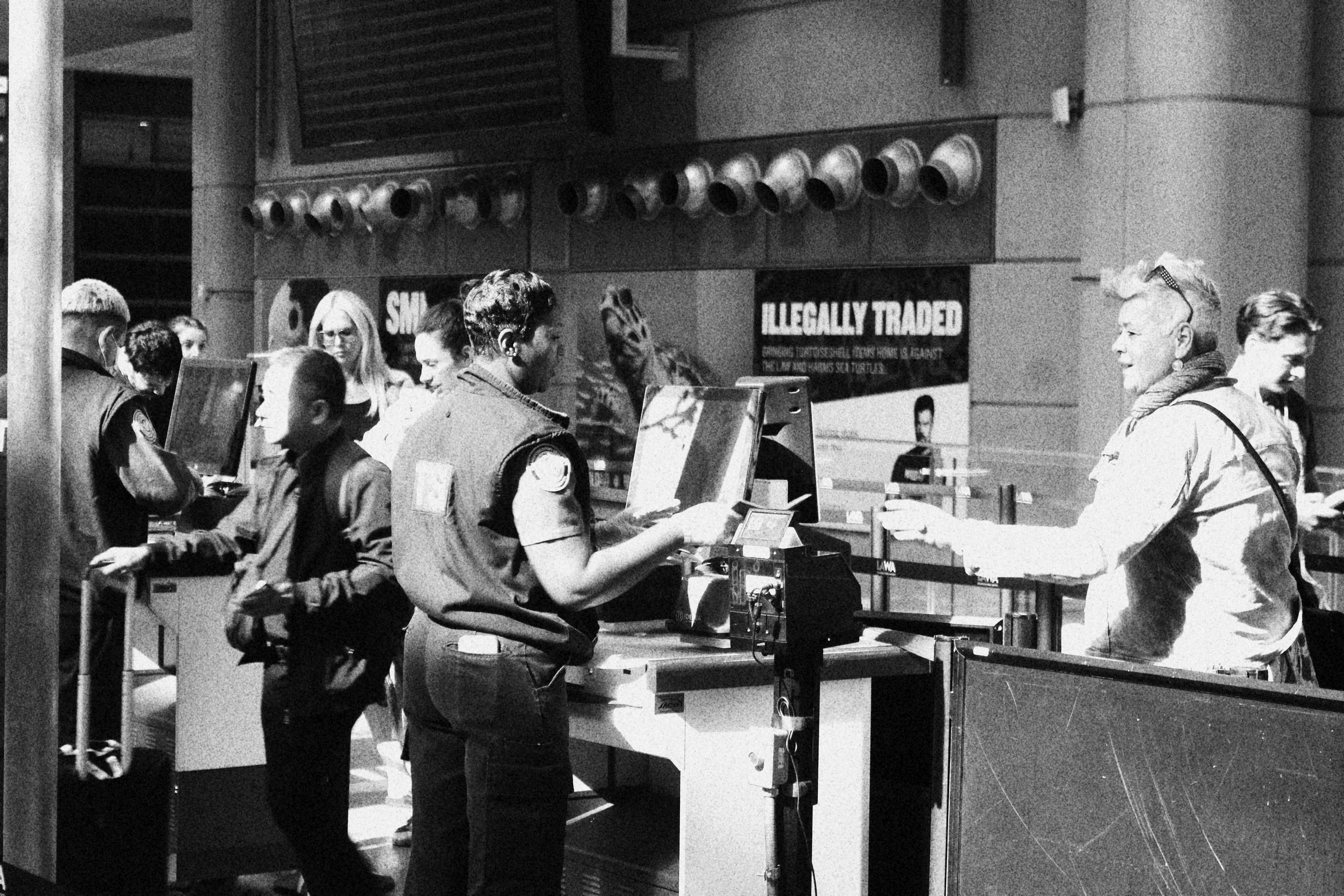 People standing in line at a transportation checkpoint, with uniformed personnel checking documents, in an indoor public space.