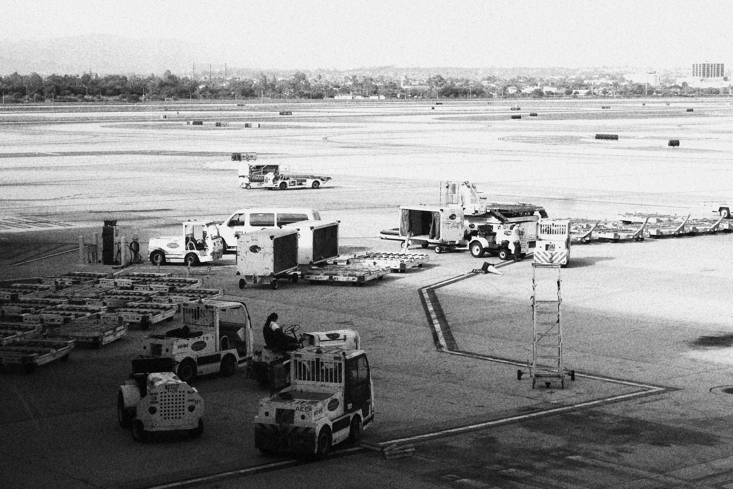 Black and white photograph of an airport tarmac with luggage carts, service vehicles, and ground crew preparing for or after a flight. In the background, taxiways and runways extend with scattered cargo containers and distant buildings.
