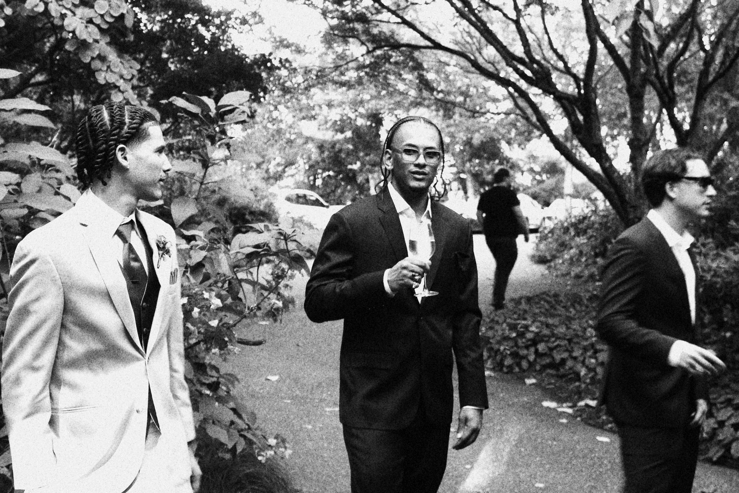 Black and white photo of three people dressed in suits, outdoors, with trees and a person walking in the background.
