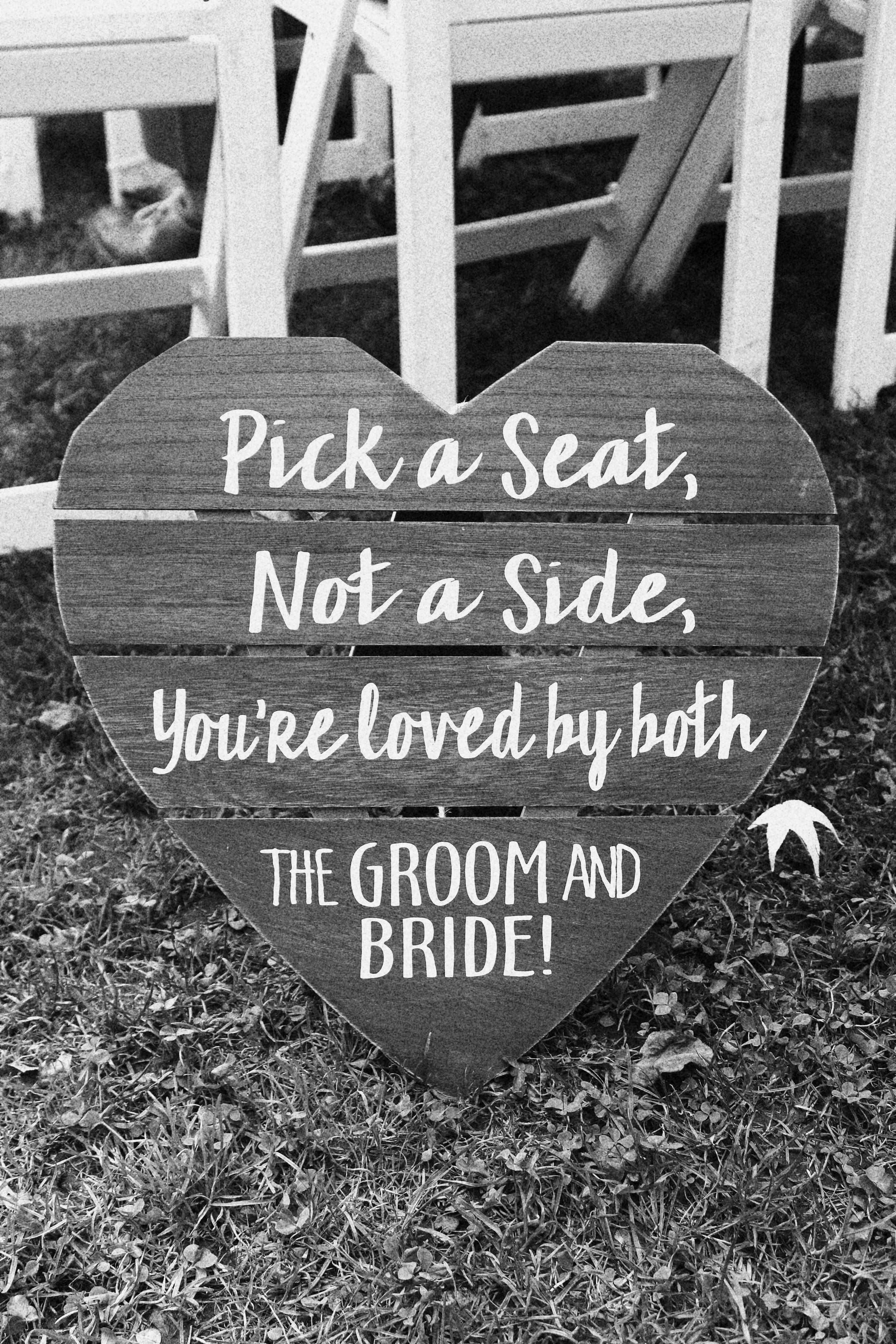 A heart-shaped wooden sign with the message: "Pick a seat, Not a Side, You're loved by both THE GROOM AND BRIDE!" placed on grass with wooden chairs in the background.