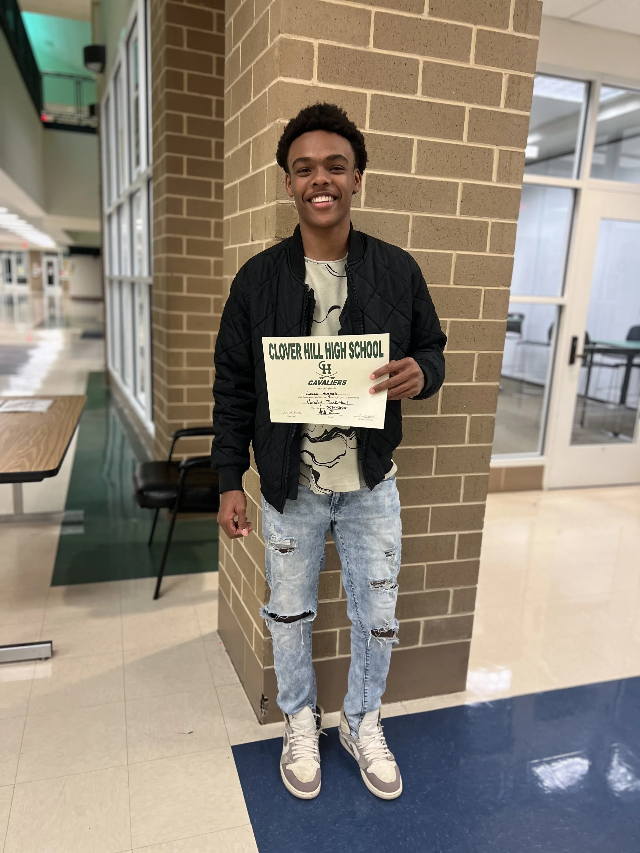A young man smiling and holding a certificate from Clover Hill High School, standing indoors against a brick wall.