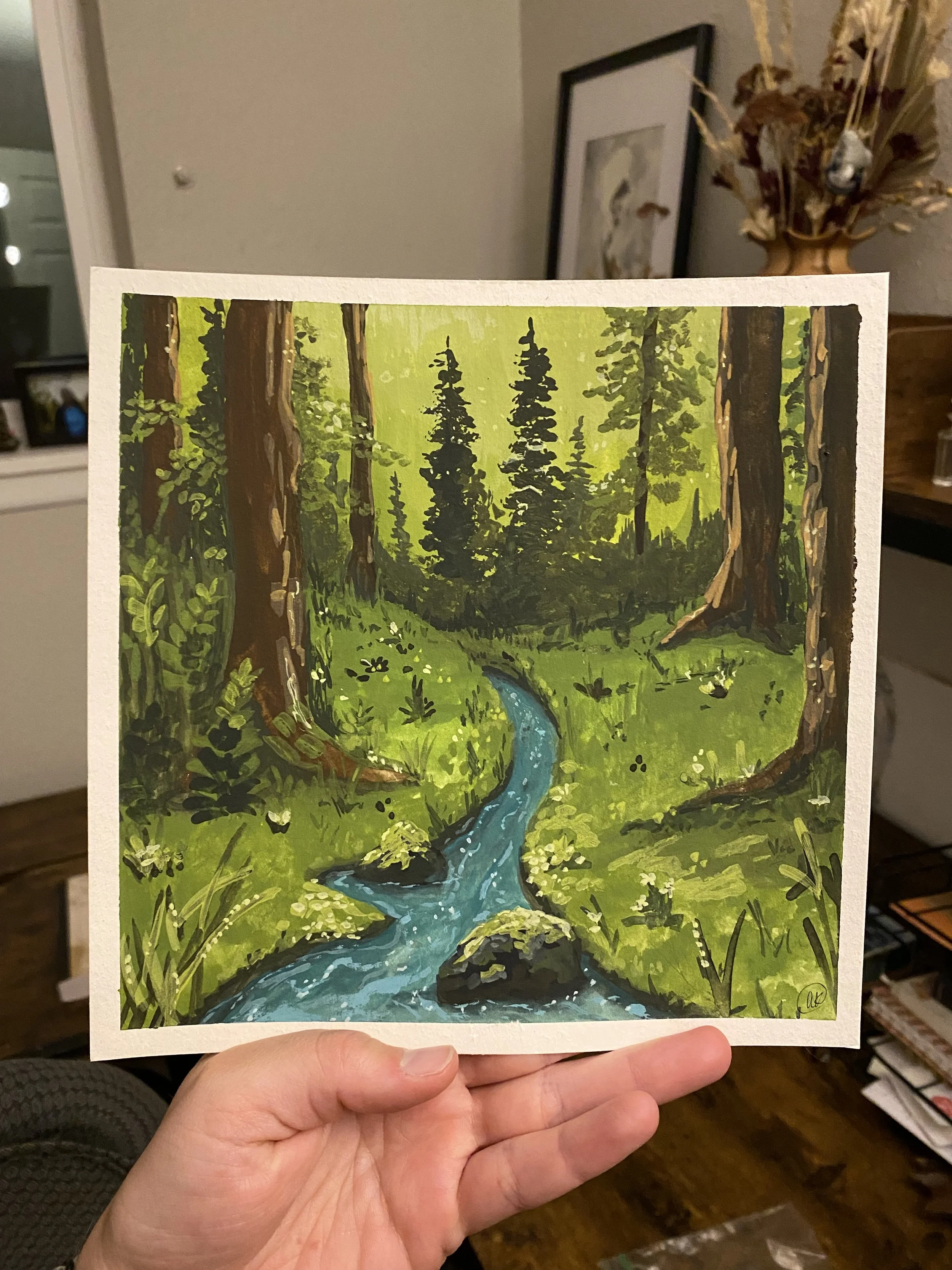 A hand holding a painting of a forest scene with tall trees, green foliage, a small stream, and rocks.