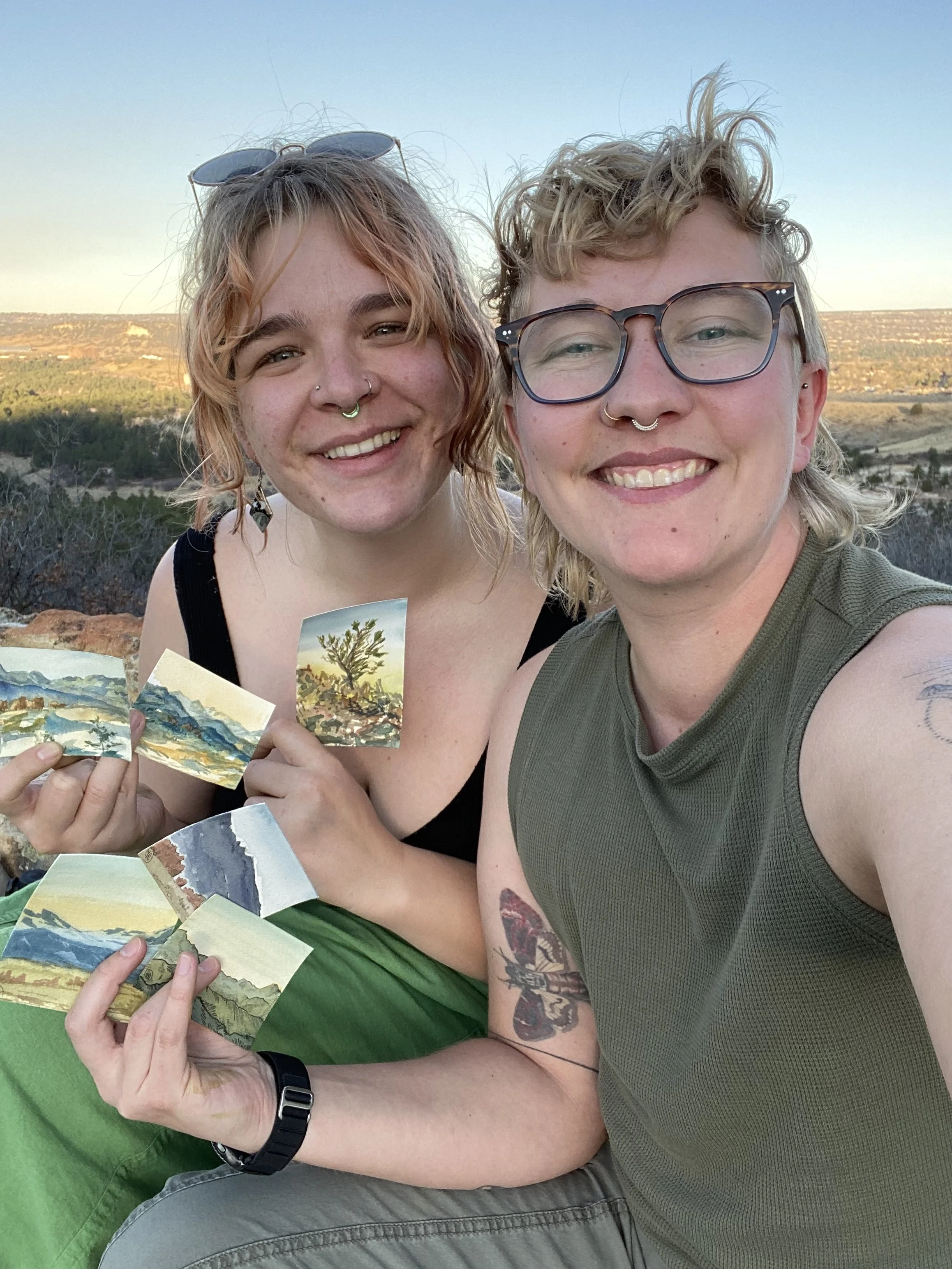 Two smiling women outdoors holding small landscape paintings, with a scenic background of rolling hills and a clear sky.