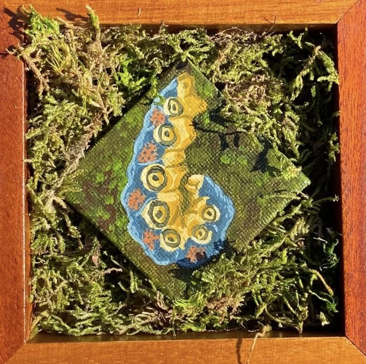 A small canvas painting of a blue butterfly with yellow and orange spots on its wings, resting on a green textured background, placed inside a wooden box filled with green moss.