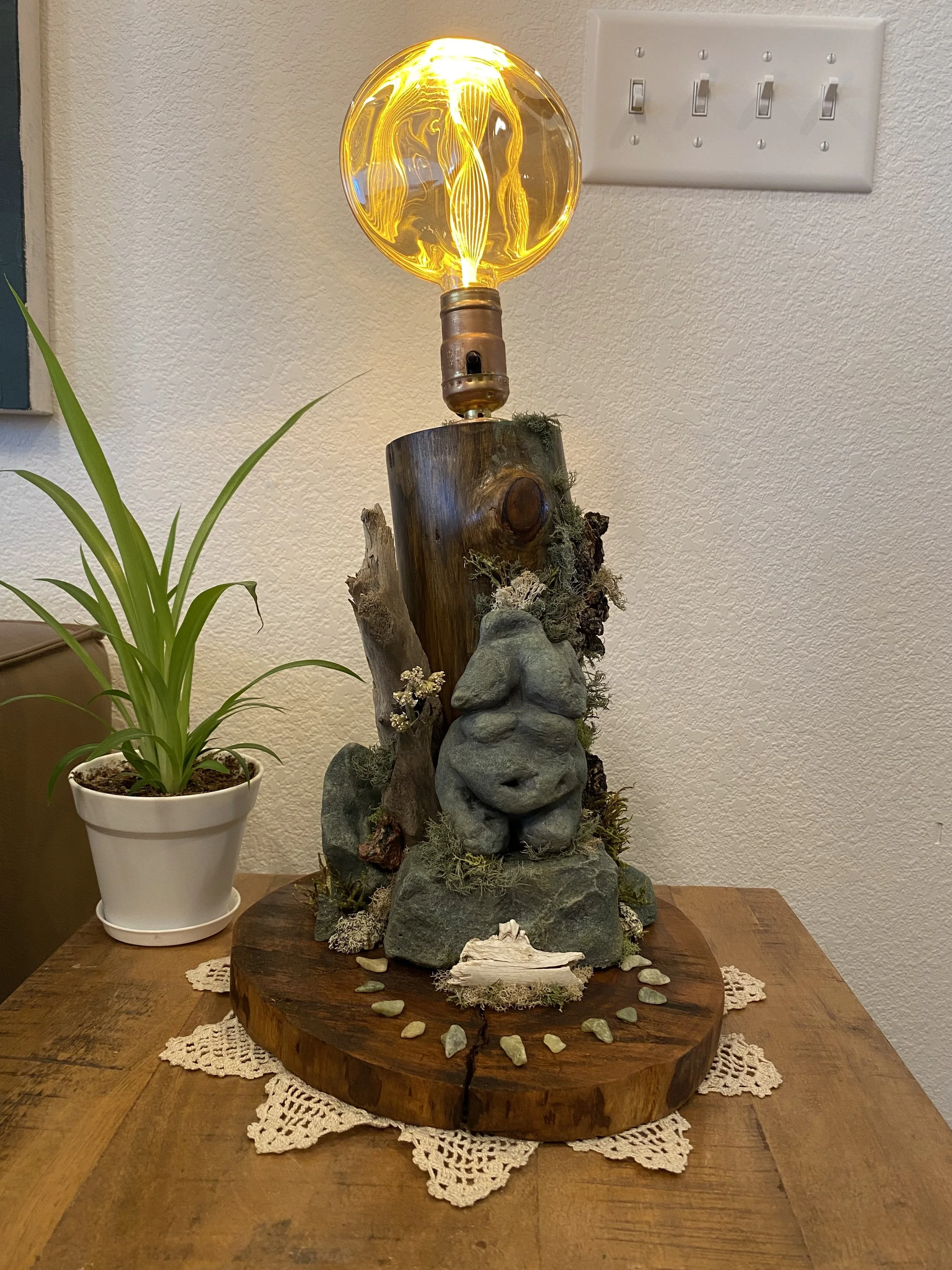 A decorative table lamp made from a large wooden log base, with a stonefigure, moss, and small rocks. The lamp has a vintage-style filament bulb on top, illuminating softly. A potted green plant is beside it on a wooden table with a lace doily underneath.