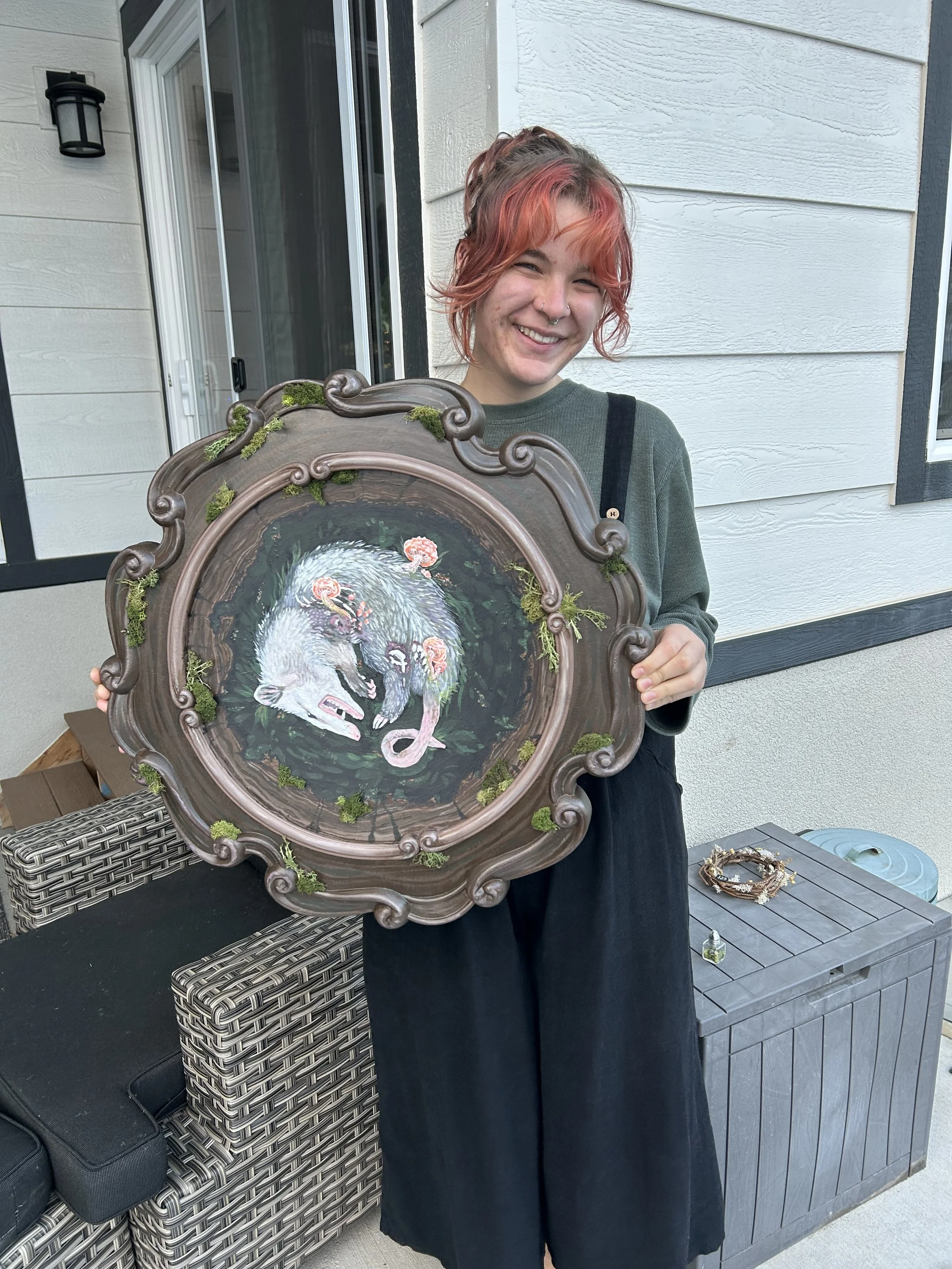 A young woman with curly pink hair and a nose piercing holding a framed painting of two white rats perched on a branch, with a dark background and pink flowers.