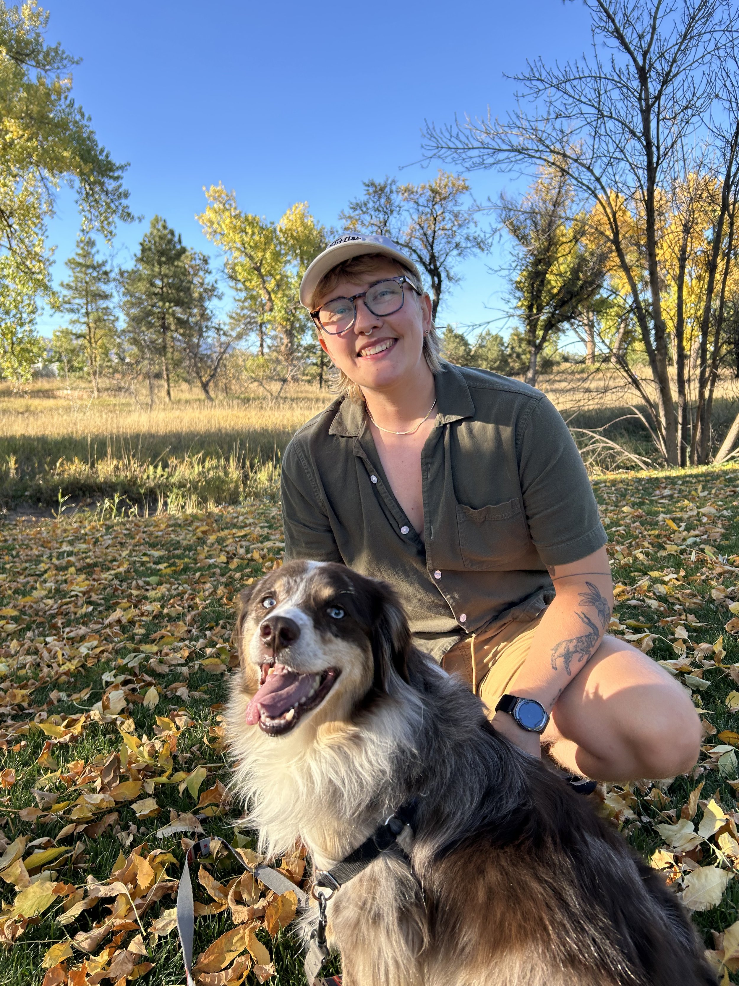 A woman with short hair, glasses, and tattoos kneeling on the ground, smiling, with a Australian Shepherd dog with blue eyes sitting in a park with yellow and green leaves, trees, and a clear blue sky.