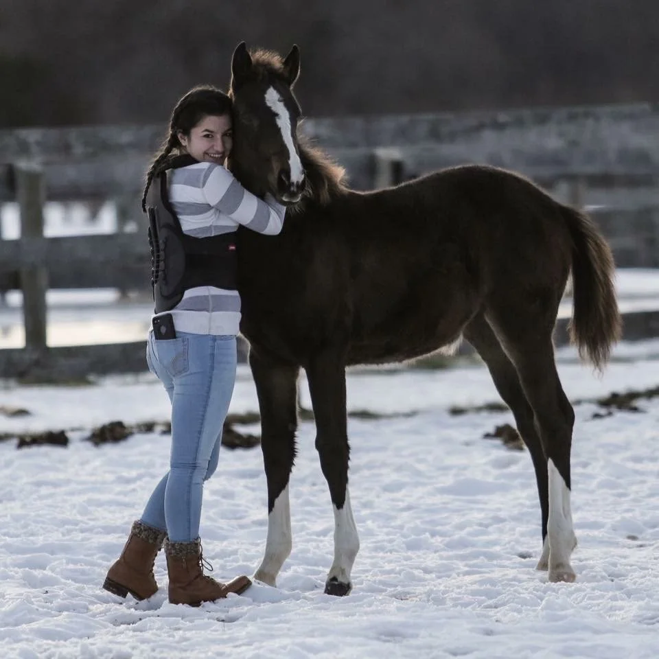A woman hugging a foal or young horse with a dark brown coat and white markings on a snowy field, smiling at the camera.