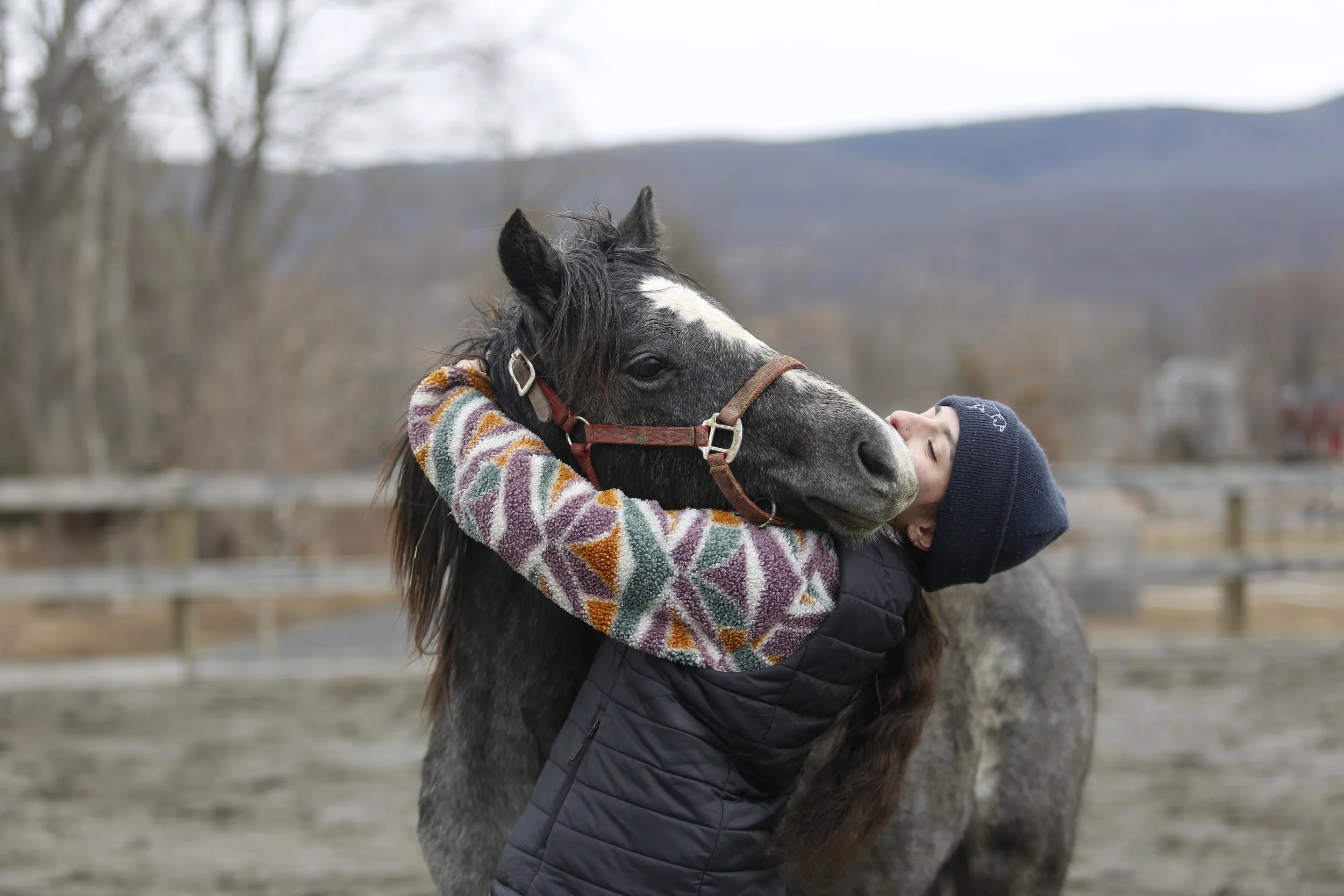A young person hugging a gray horse with a white stripe on its face outdoors, with trees and mountains in the background, during a chilly day.