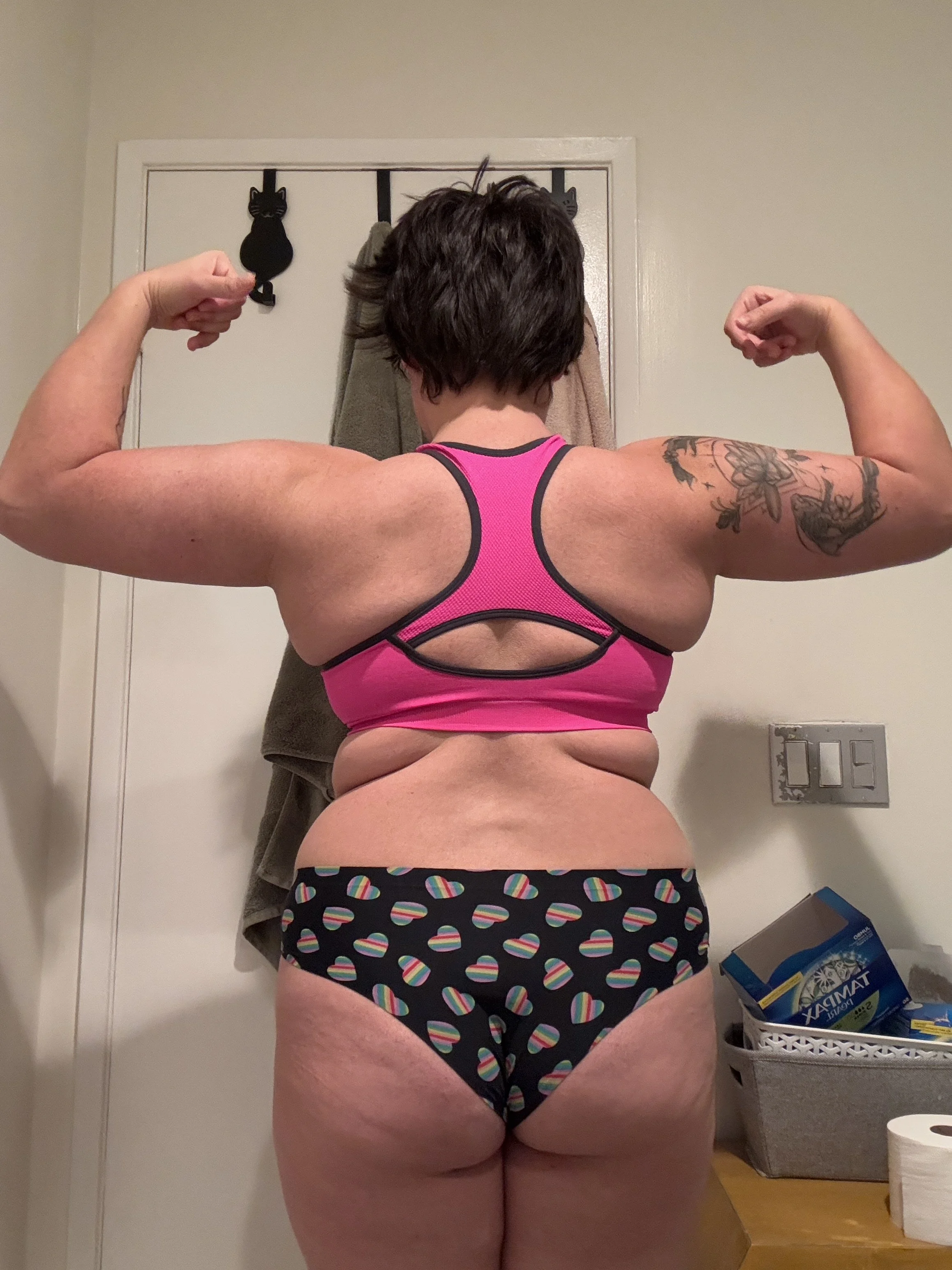 A woman with short dark hair flexing her arms in a mirror. She is wearing a pink sports bra and black shorts with rainbow shades. Her back has a tattoo on her left arm. The background shows a towel hanging on a door and a basket with various items.