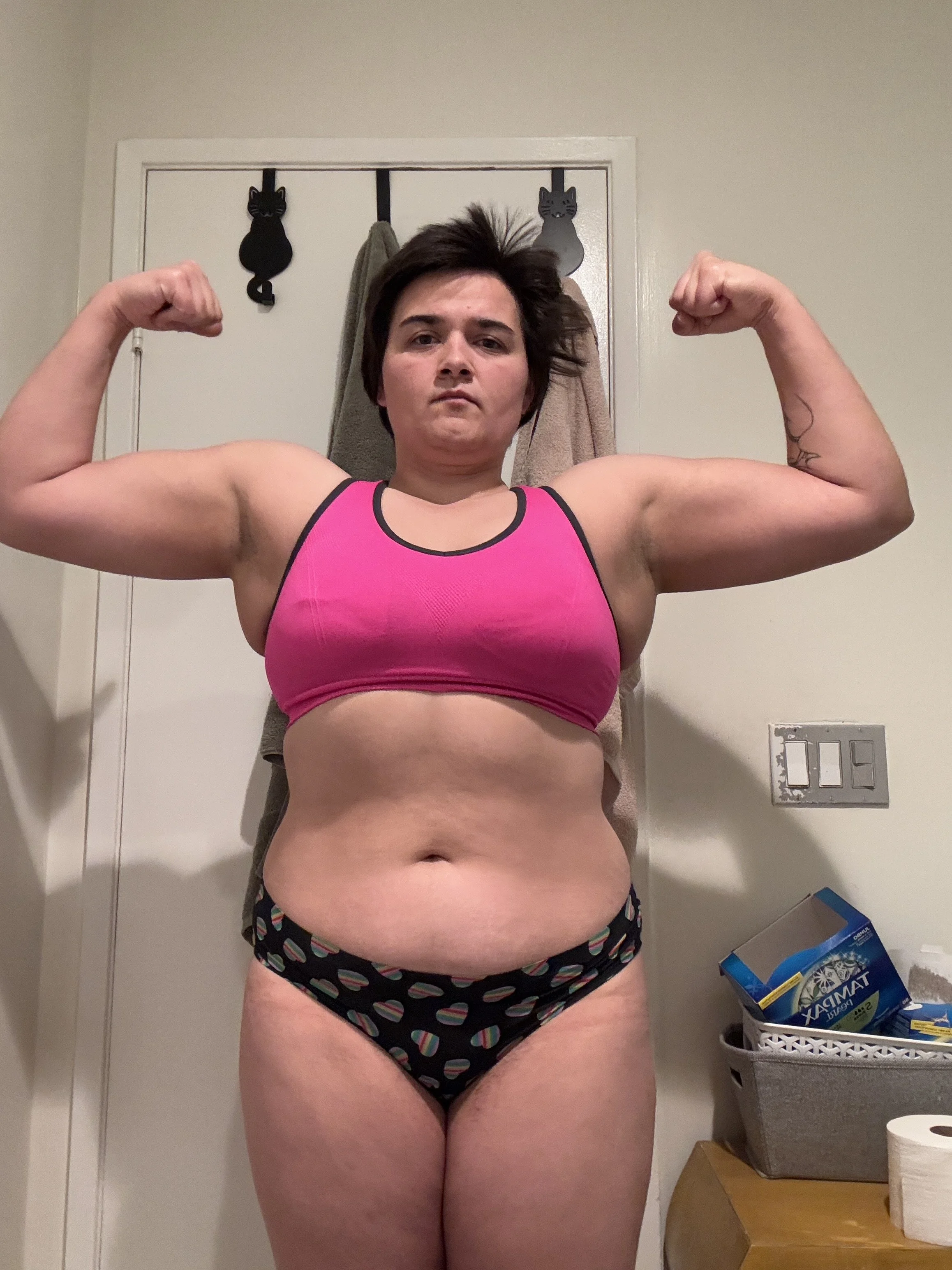 A woman with short dark hair flexing her biceps in a mirror, wearing a bright pink sports bra and patterned underwear, standing in a room with a white door and towels hanging behind her.