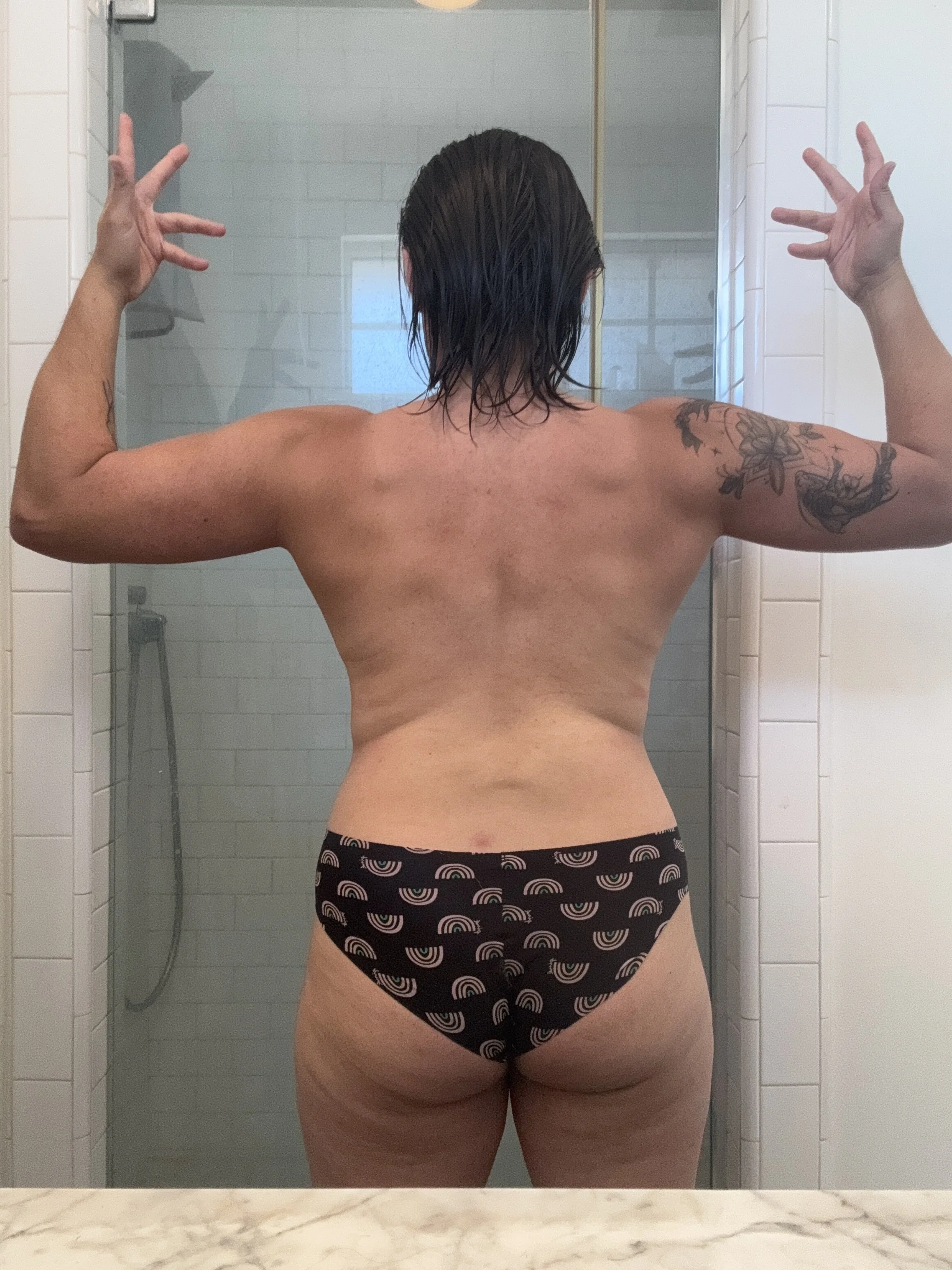 Back view of a person flexing their muscles in a bathroom mirror, wearing black underwear with rainbow arches, with tattoos on both arms and short wet hair.