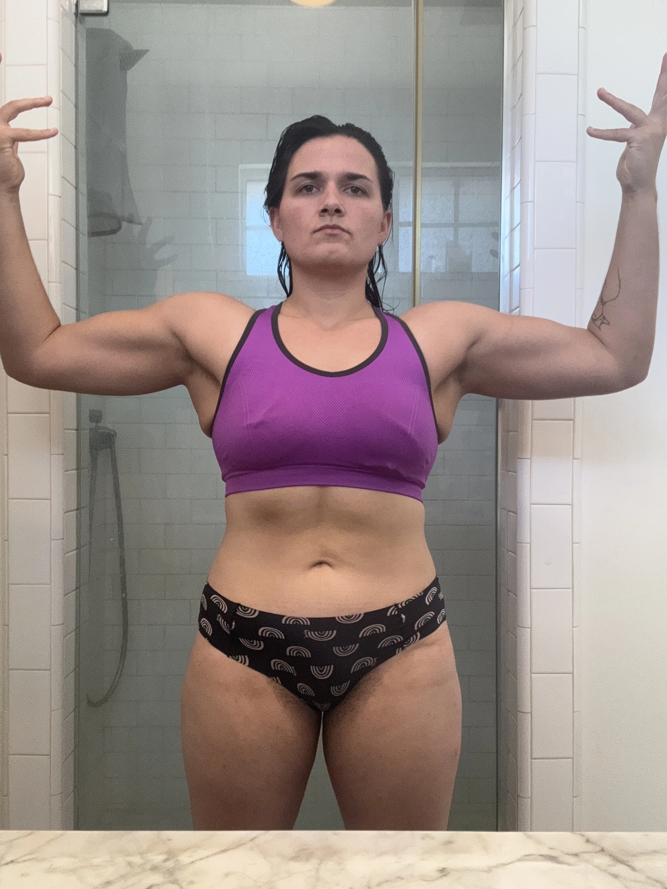 Woman with dark hair in a purple sports bra and black underwear with rainbow patterns, flexing her muscles in front of a bathroom mirror.