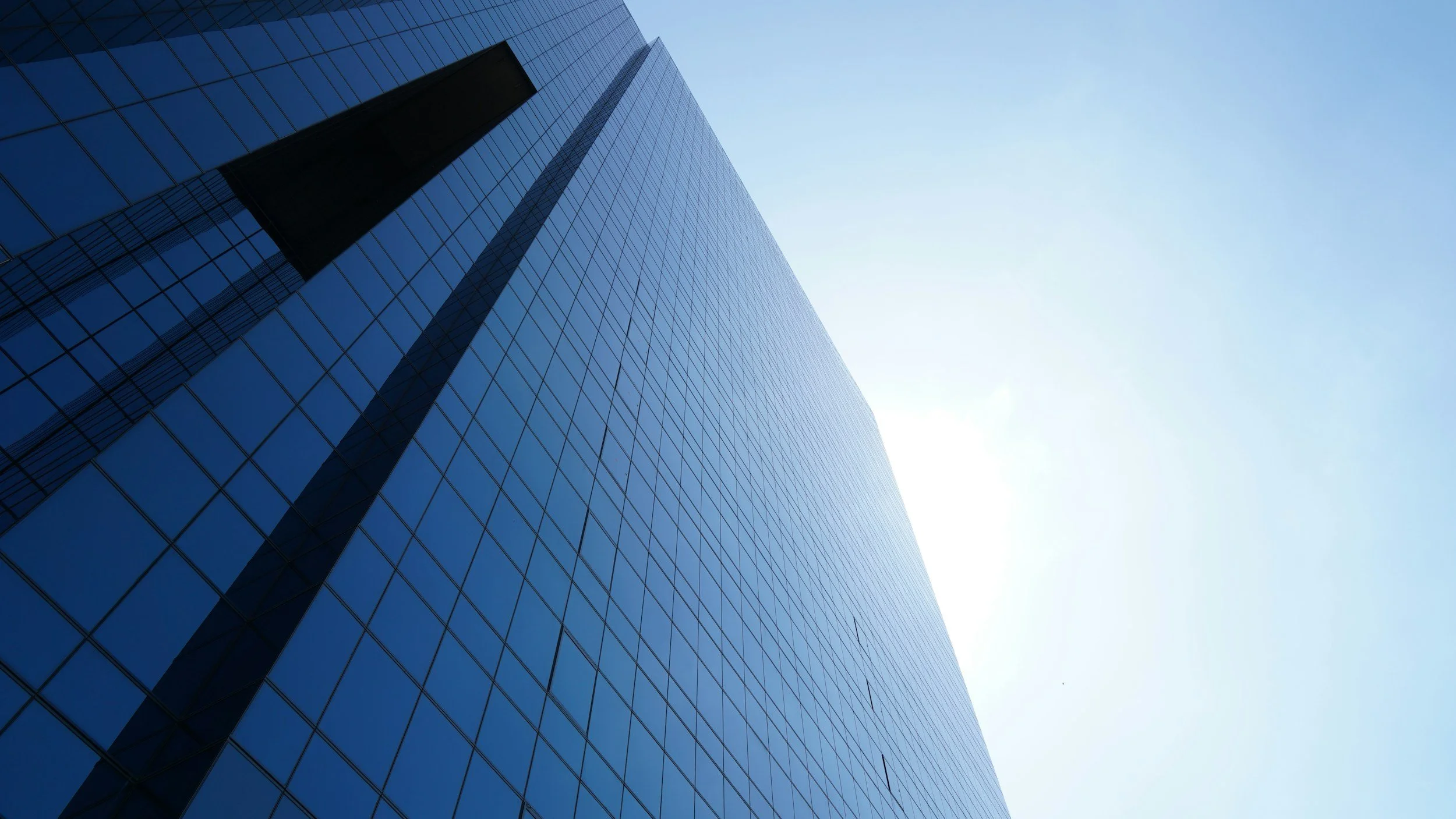 A modern glass skyscraper with a reflective surface, shot from below against a clear blue sky.