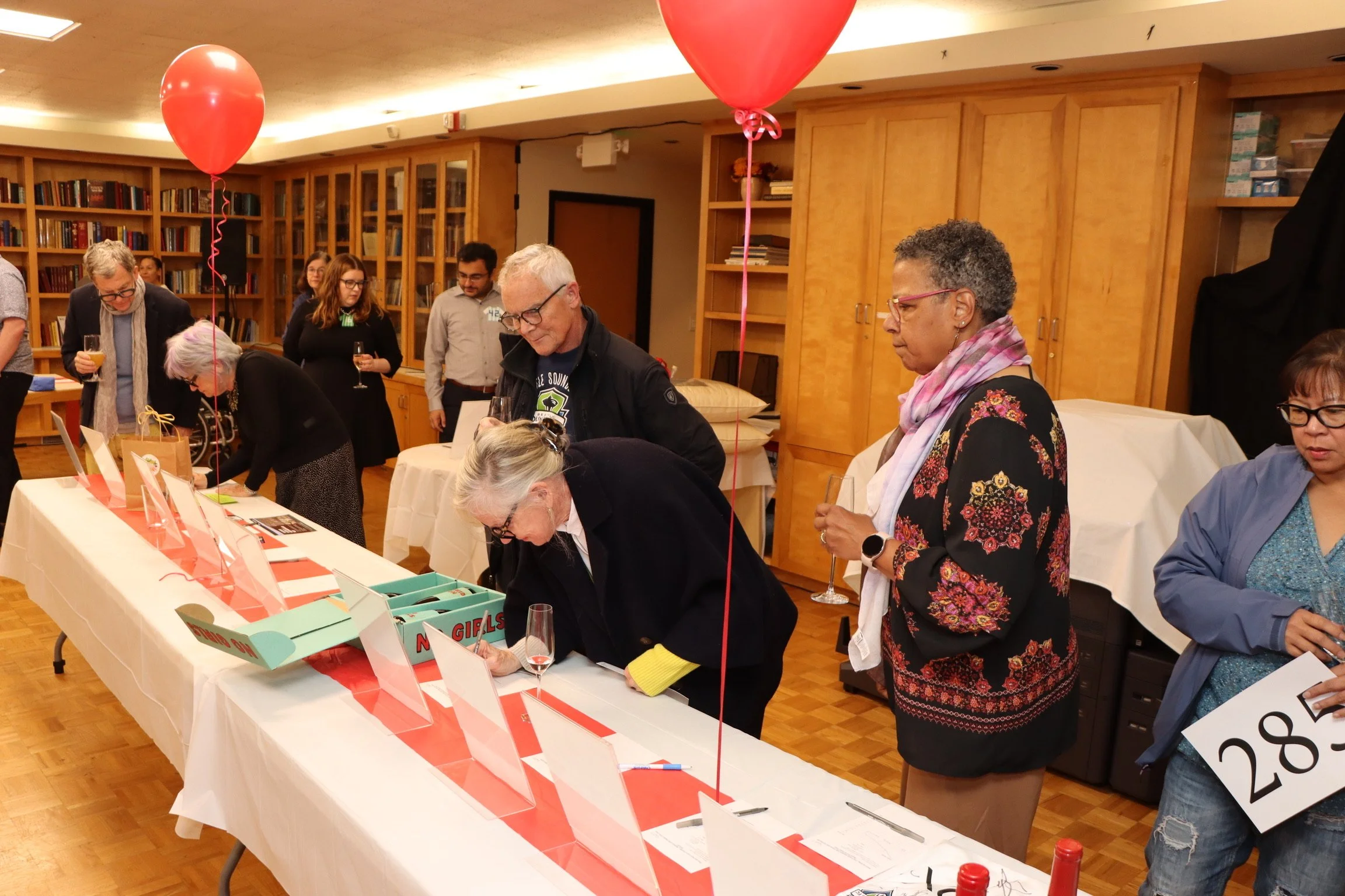 People gathered around a table with pink balloons, at a social event in a room with wooden bookshelves and cabinets, some holding glasses of drinks.