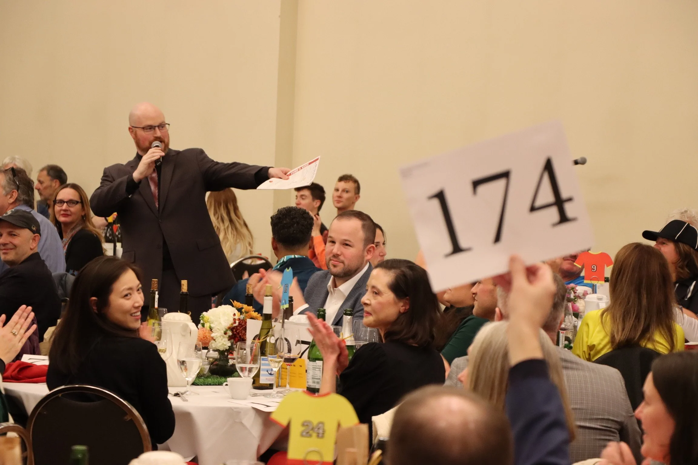 A man in a suit holding a microphone and a paper, speaking at a large banquet or event. The room is filled with seated people, some clapping, with tables decorated with flowers, wine bottles, and tableware. There are signs with the number 174 and people wearing casual and semi-formal attire.