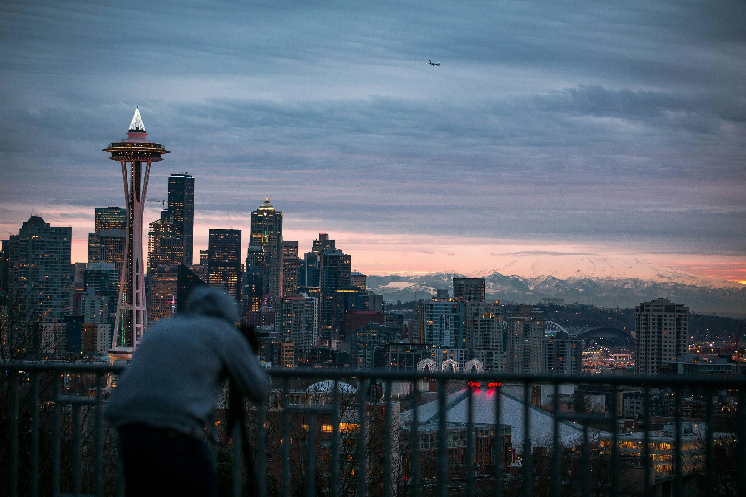 City skyline of Seattle with the Space Needle and high-rise buildings, overcast sky, mountains in the distance, and a plane flying overhead. In the foreground, a person with a hood is visible, standing behind a railing.