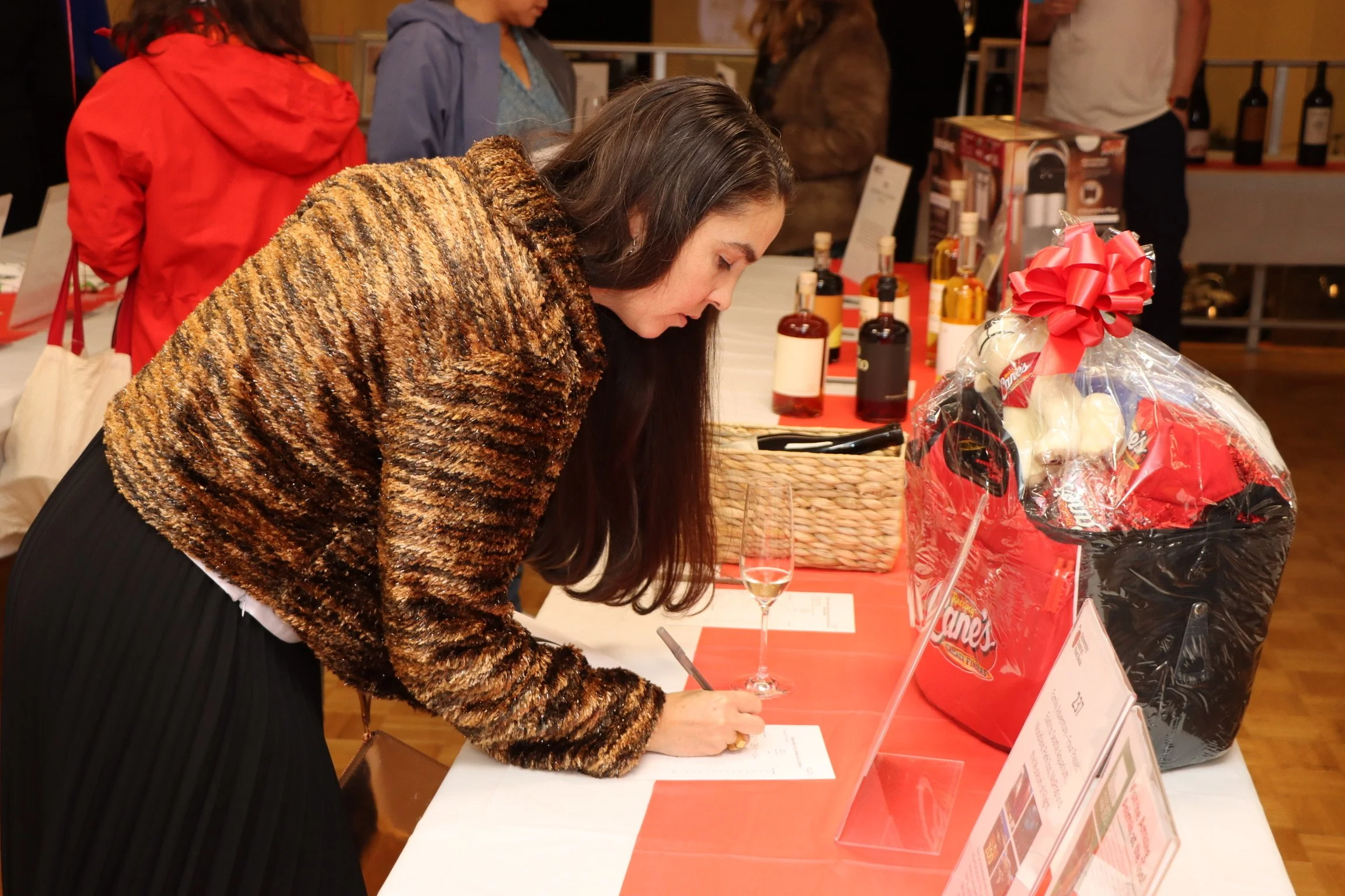 Woman with long dark hair dressed in a leopard print jacket signing a document at a table during an event. The table displays gift baskets, gift bags, and informational flyers. Other attendees are visible in the background.