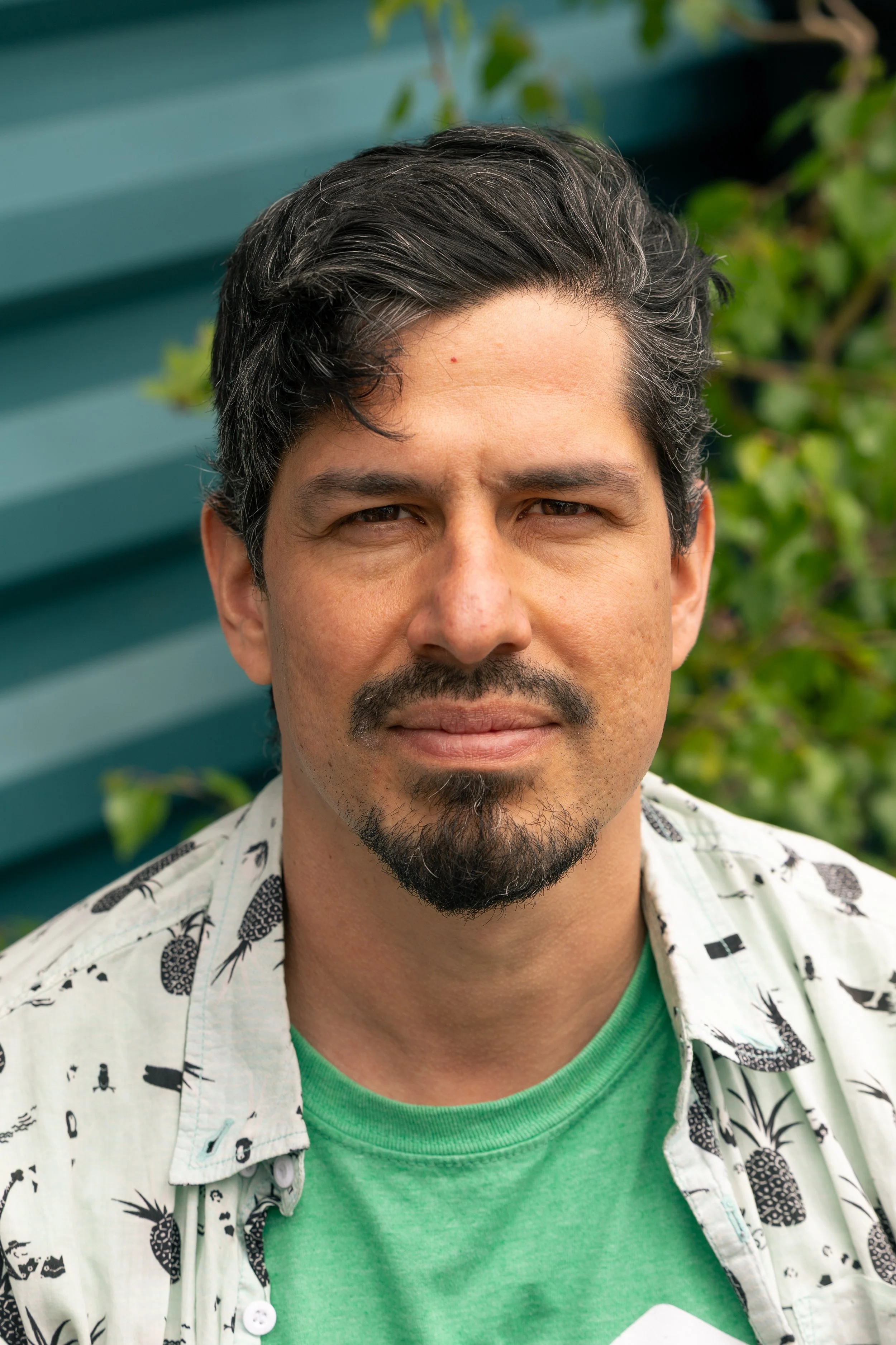 A man with dark, wavy hair, a goatee, and mustache, wearing a green shirt underneath a white short-sleeved button-up shirt with a black leaf pattern, standing outdoors in front of a green wooden fence with foliage in the background.