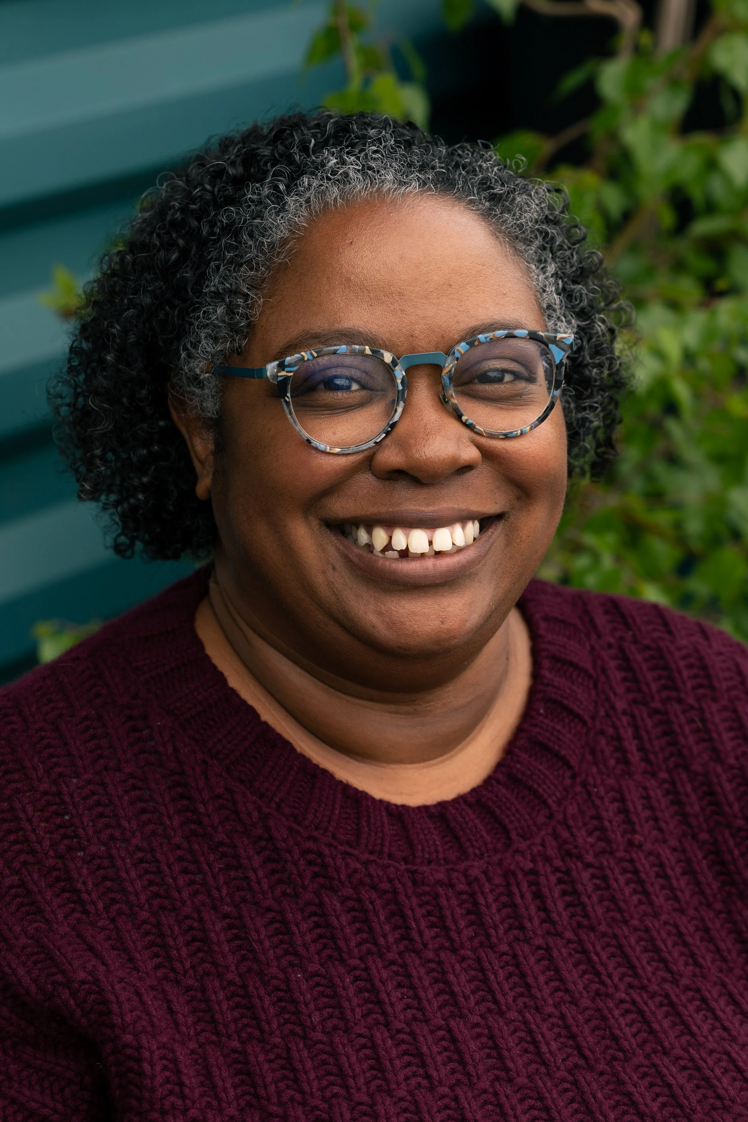 A smiling woman with curly salt-and-pepper hair and patterned glasses, wearing a maroon sweater, standing outdoors with green foliage and a teal-colored background.