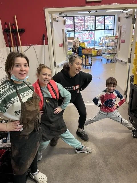 Four children and an adult are posing in a nursery or classroom. The children are smiling, and the adult is making a playful pose. In the background, a woman is setting up or organizing supplies near a window with a view outside.