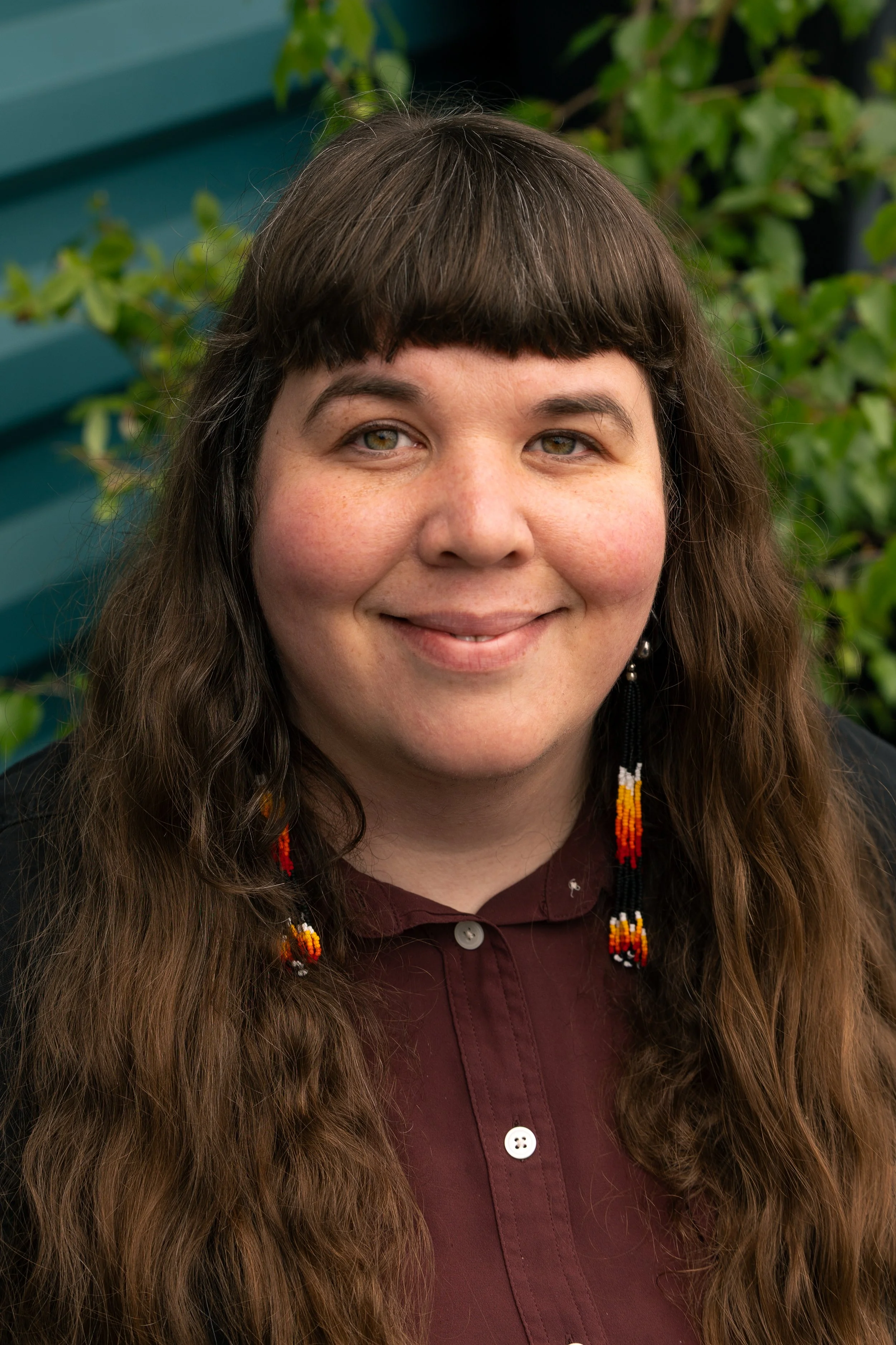 Close-up of a woman with long brown hair, wearing beaded earrings and a maroon button-up shirt, smiling outdoors with green foliage in the background.