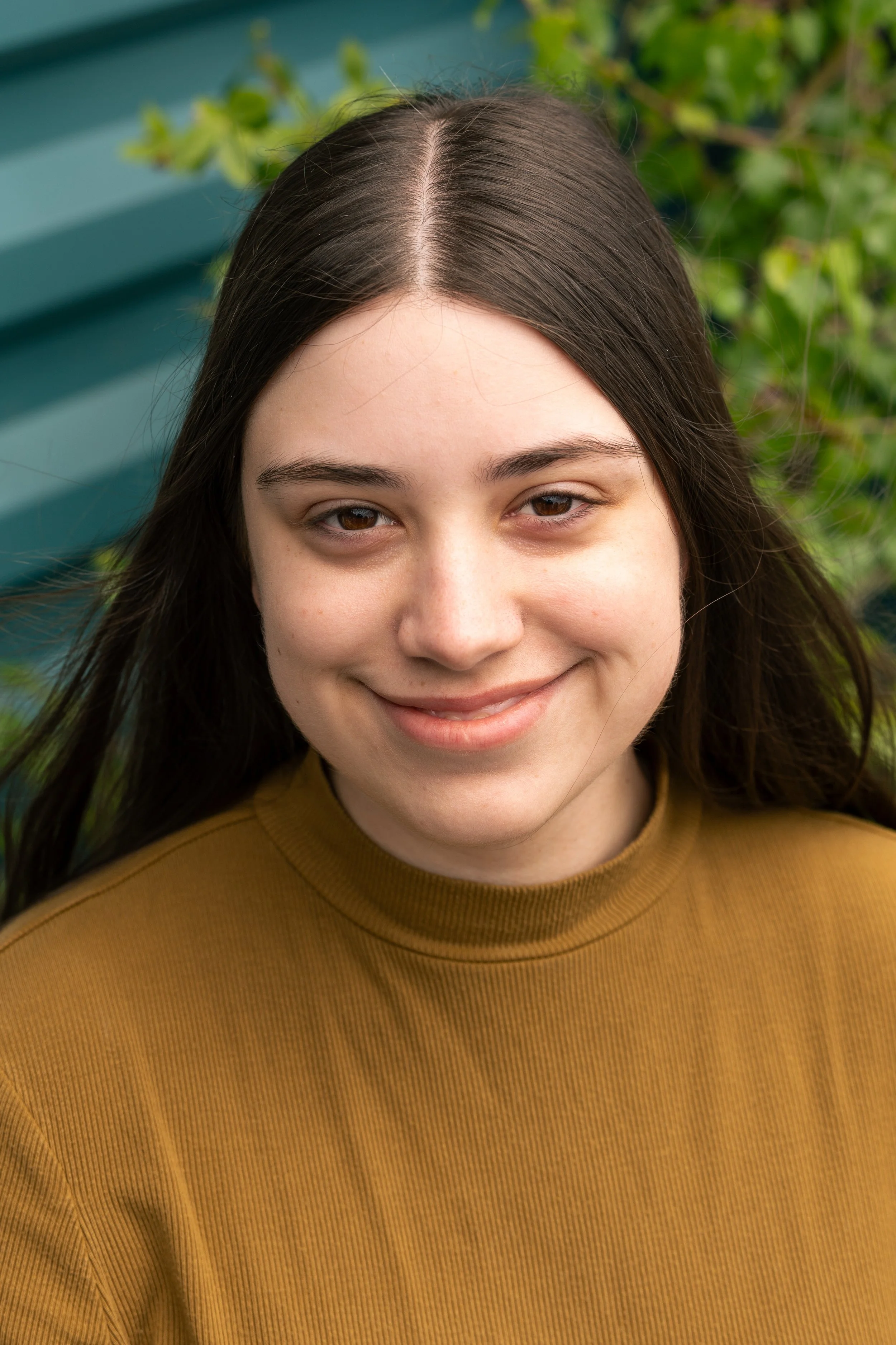Close-up of a young woman with long dark hair, smiling, wearing a mustard-colored top, outdoors with green foliage and a teal wooden structure in the background.